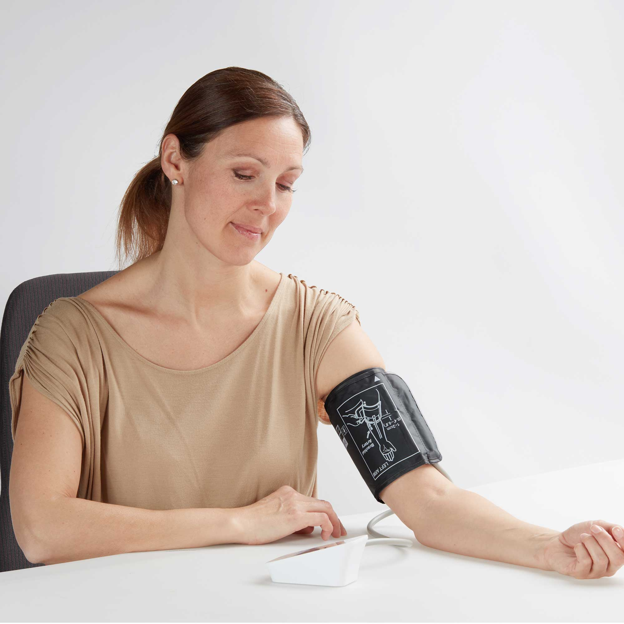 Frau misst Blutdruck mit einem Gerät am Arm. Sie sitzt an einem weißen Tisch.