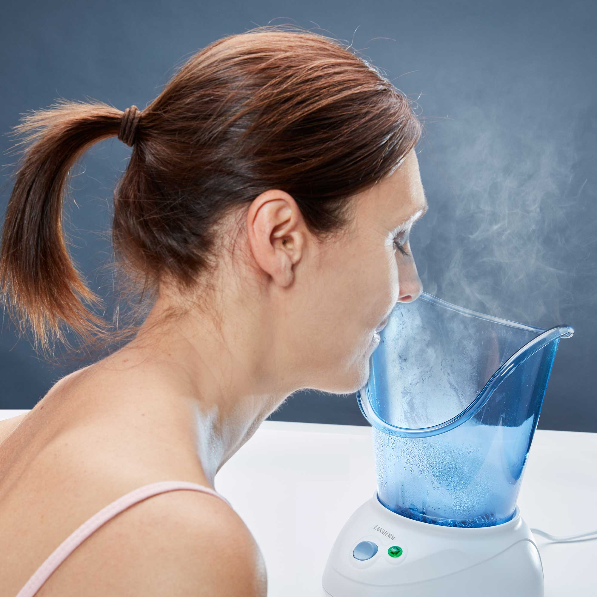 Een vrouw die haar gezicht stoomt met een gezichtsstomer, die blauw en wit is, op een witte tafel.