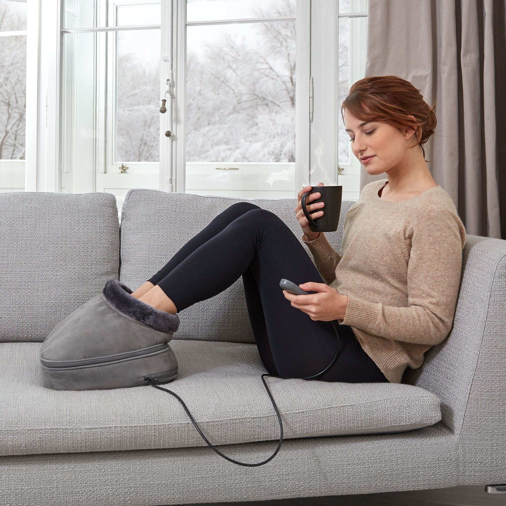 Woman sitting on a couch with a heated foot warmer and a mug, looking down at a remote.