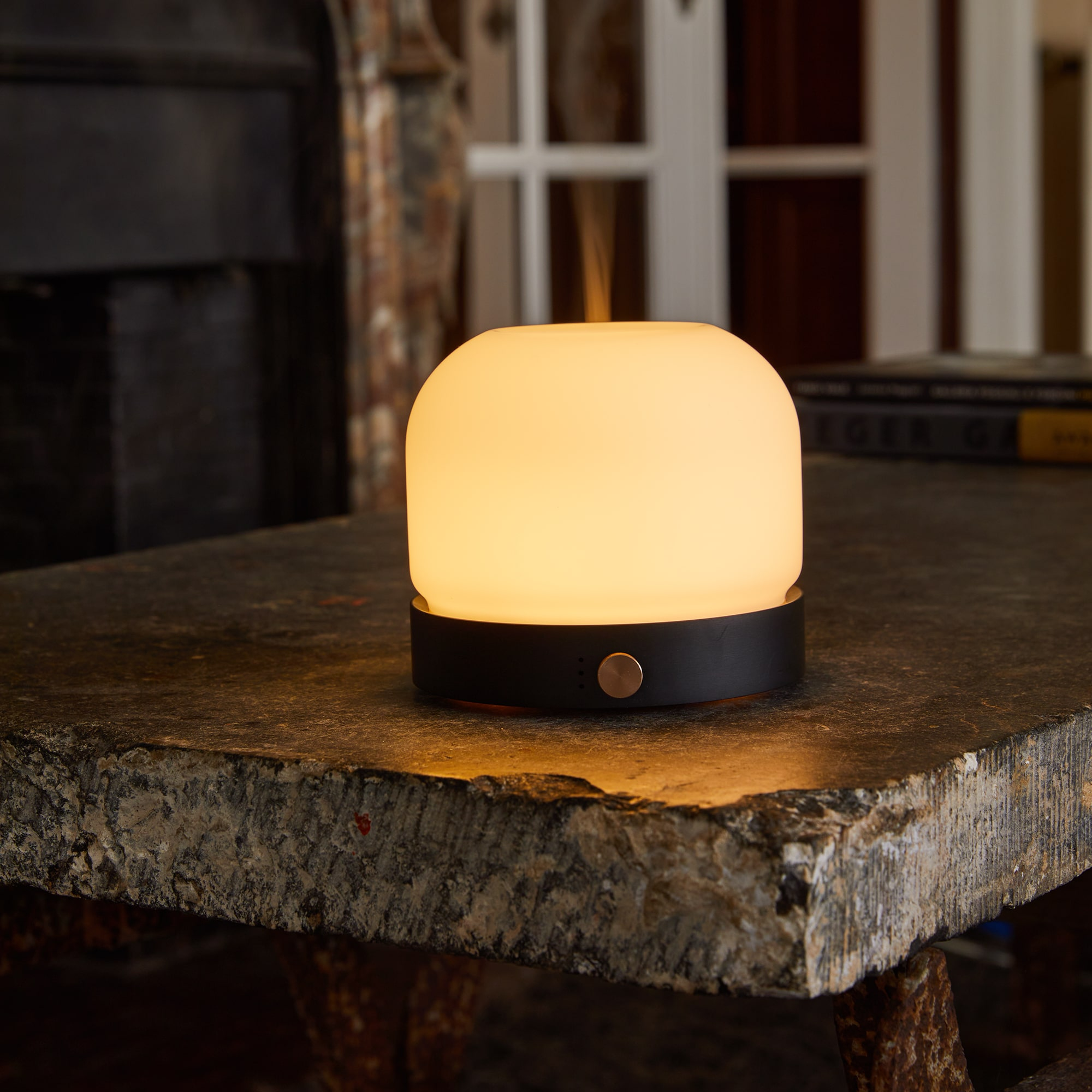 White and black lamp on a table. It is glowing. The background is a fireplace.