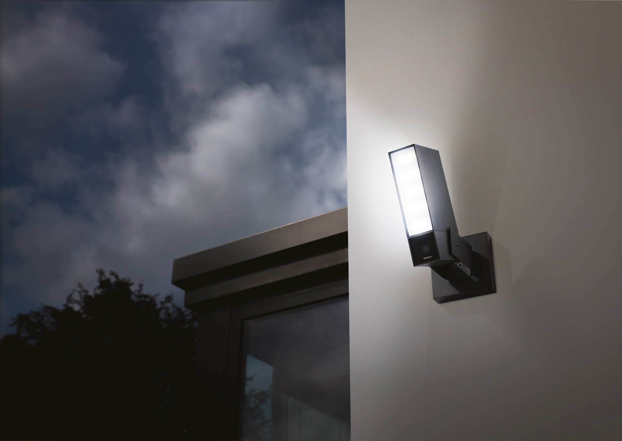 Luz de seguridad negra en una pared al anochecer. La luz está encendida, iluminando la pared. Cielo nublado.