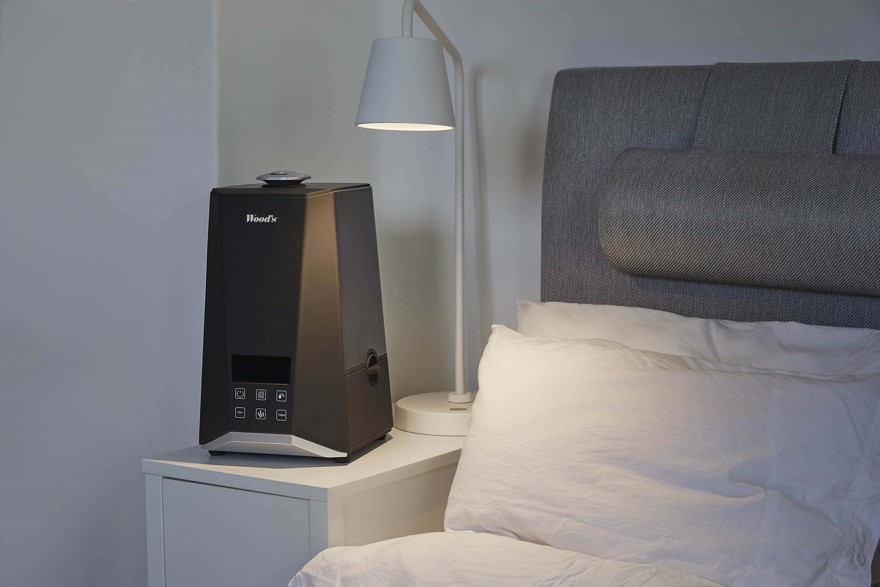 Un umidificatore nero su un comodino bianco con una lampada bianca e un letto con cuscini sullo sfondo.