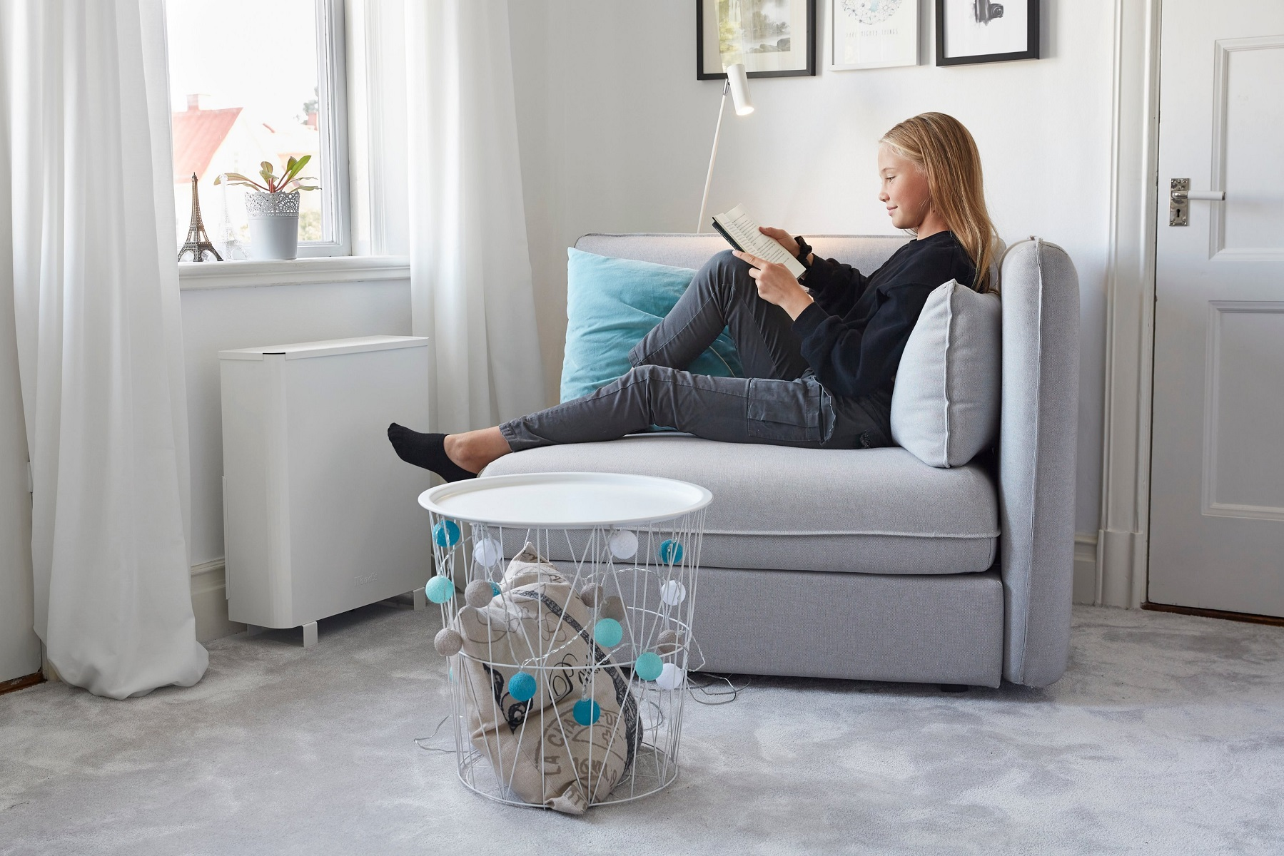Mädchen liest auf einem grauen Sofa in Fensternähe. Ein weißer Drahtkorb mit Spielzeug steht davor.