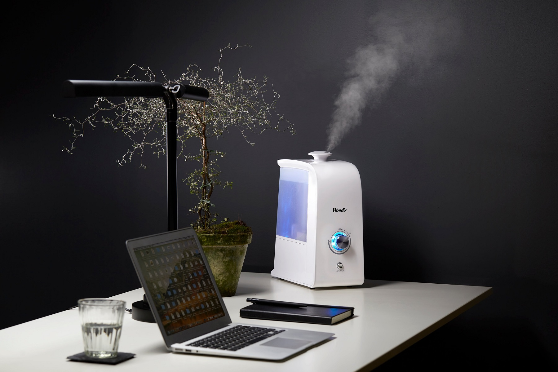 Ein Schreibtisch mit Laptop, Luftbefeuchter, Pflanze, Glas Wasser und Notizblock.