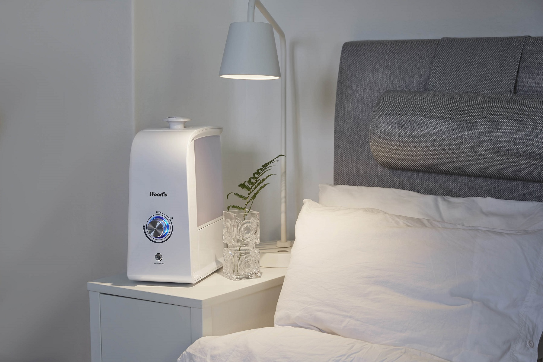 Ein weißer Luftbefeuchter auf einem Nachttisch. Eine Lampe, ein Farn und ein Bett im Hintergrund.