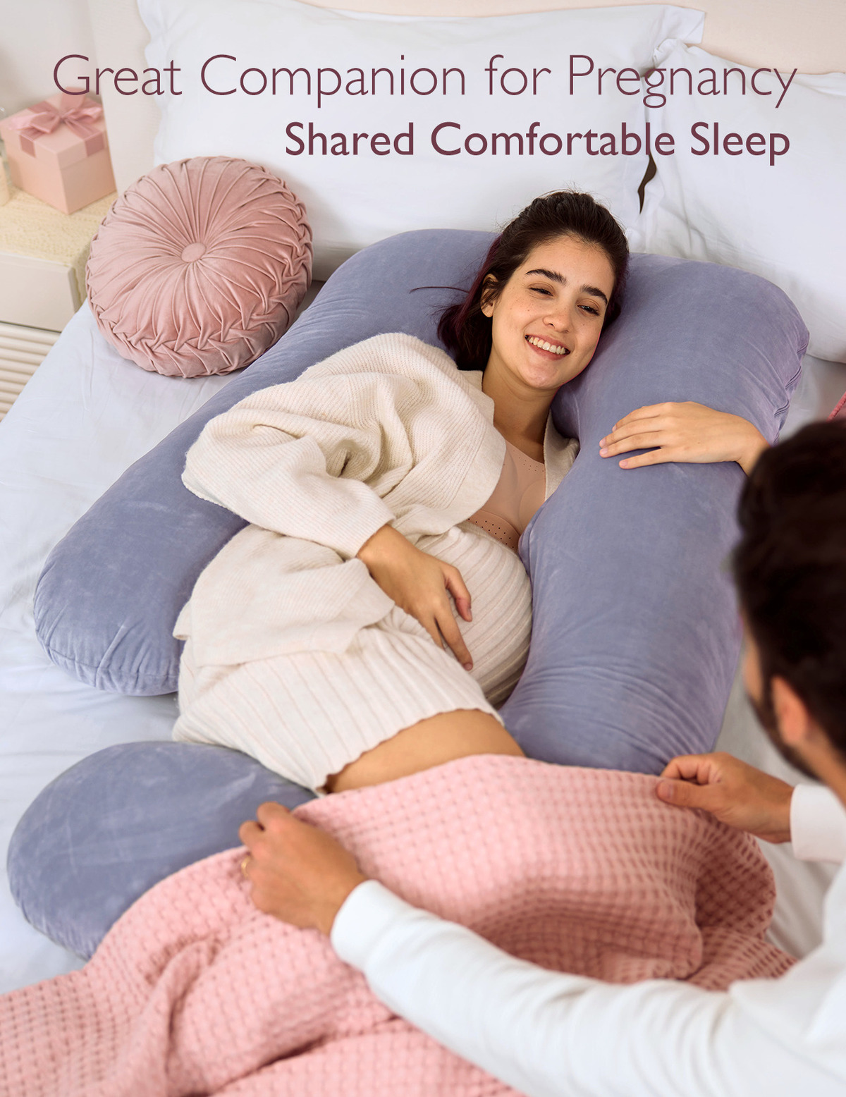 Schwangere Frau und Mann lächeln, mit einem U-förmigen Kissen auf einem Bett. Eine rosa Decke ist sichtbar.