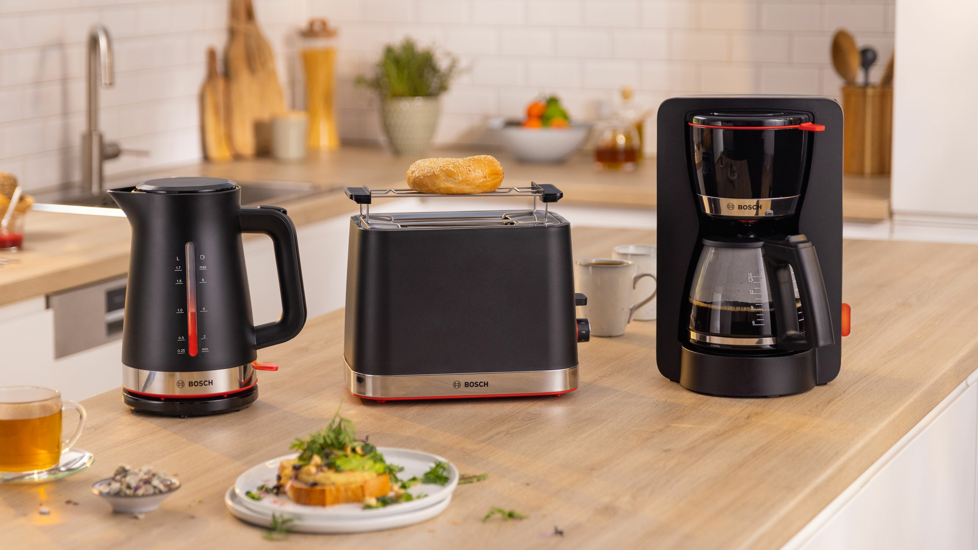 Keukenapparatuur: waterkoker, broodrooster met bagel, koffiezetapparaat en toast. Houten aanrecht. Achtergrond met raam en planten.