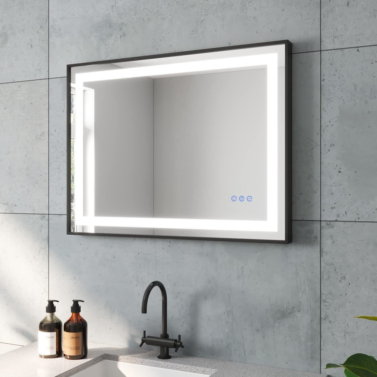 Ein rechteckiger Spiegel mit Hintergrundbeleuchtung, schwarzer Rahmen, an einer grauen Wand. Wasserhahn, Seifenflaschen und Pflanze sind zu sehen.
