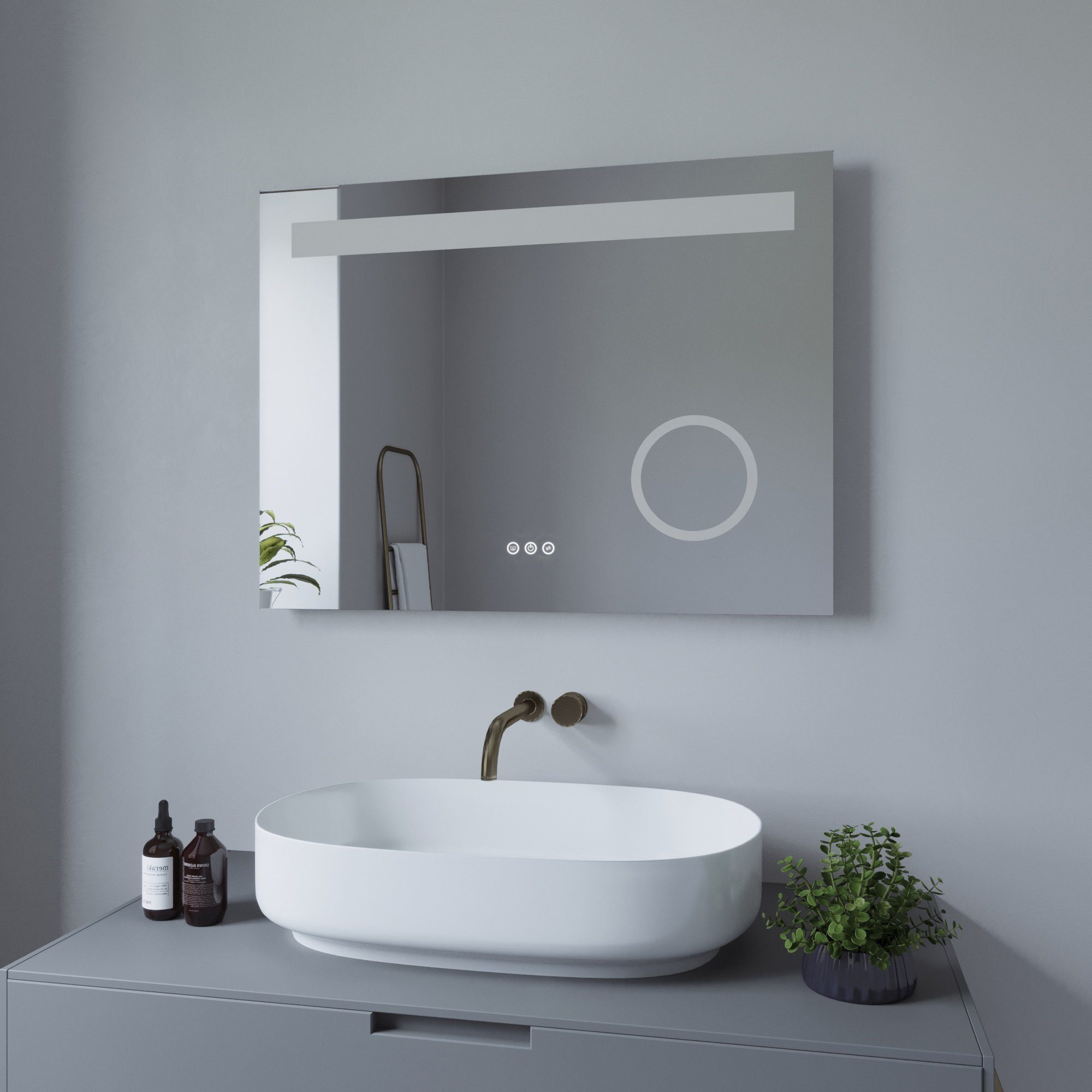 Ein beleuchteter Spiegel an einer grauen Wand, mit einem Waschbecken, Wasserhahn und einer kleinen Pflanze.