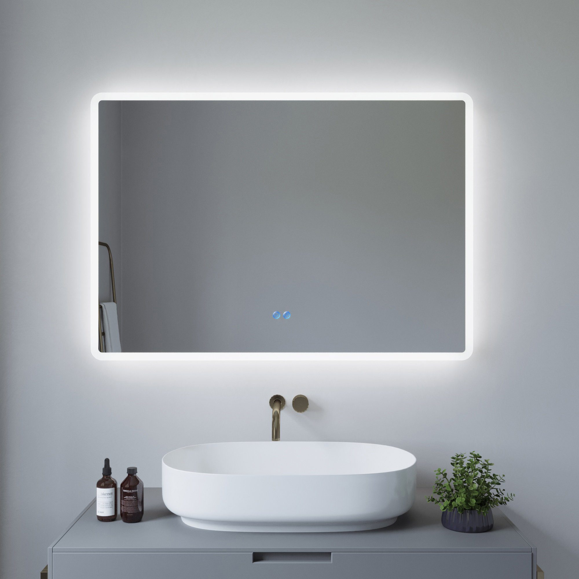 Ein rechteckiger Spiegel mit Hintergrundbeleuchtung und weißem Rahmen an einer grauen Wand. Waschbecken, Wasserhahn, Flaschen und Pflanze sind zu sehen.