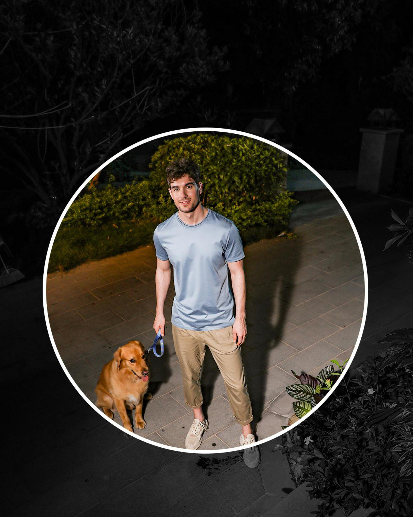 Mężczyzna spacerujący z psem w nocy, oświetlony okrągłym światłem. Ma na sobie niebieską koszulę.