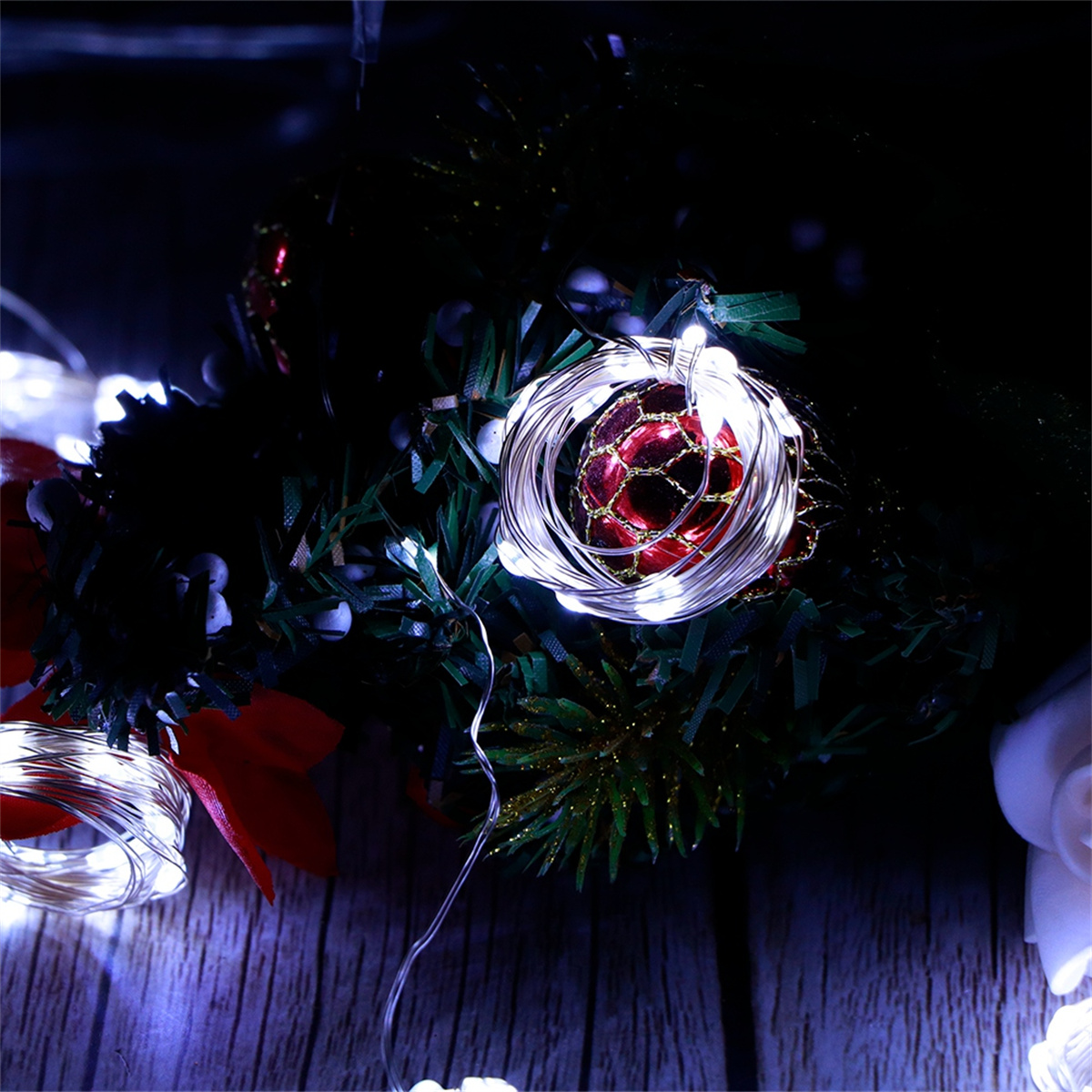 Weihnachtsbeleuchtung um roten Schmuck, auf dunkler Holzoberfläche. Grüne Tannenzweige sind sichtbar.