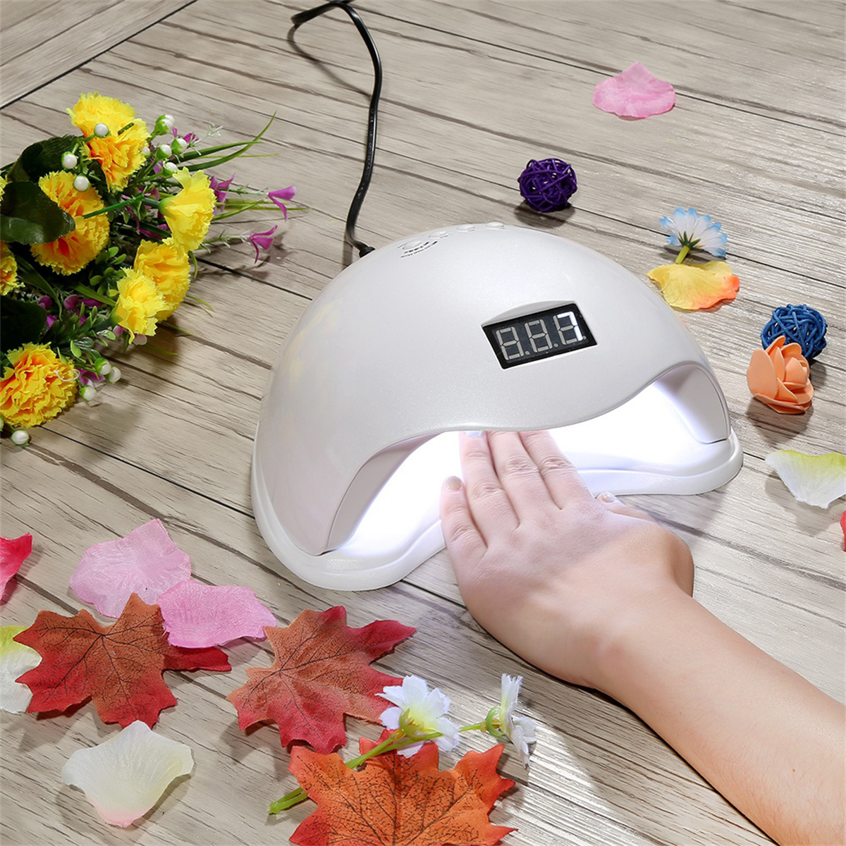Hand benutzt eine weiße UV-Nagellampe auf einem Holztisch mit Blumen und Nagelkunst-Zubehör.