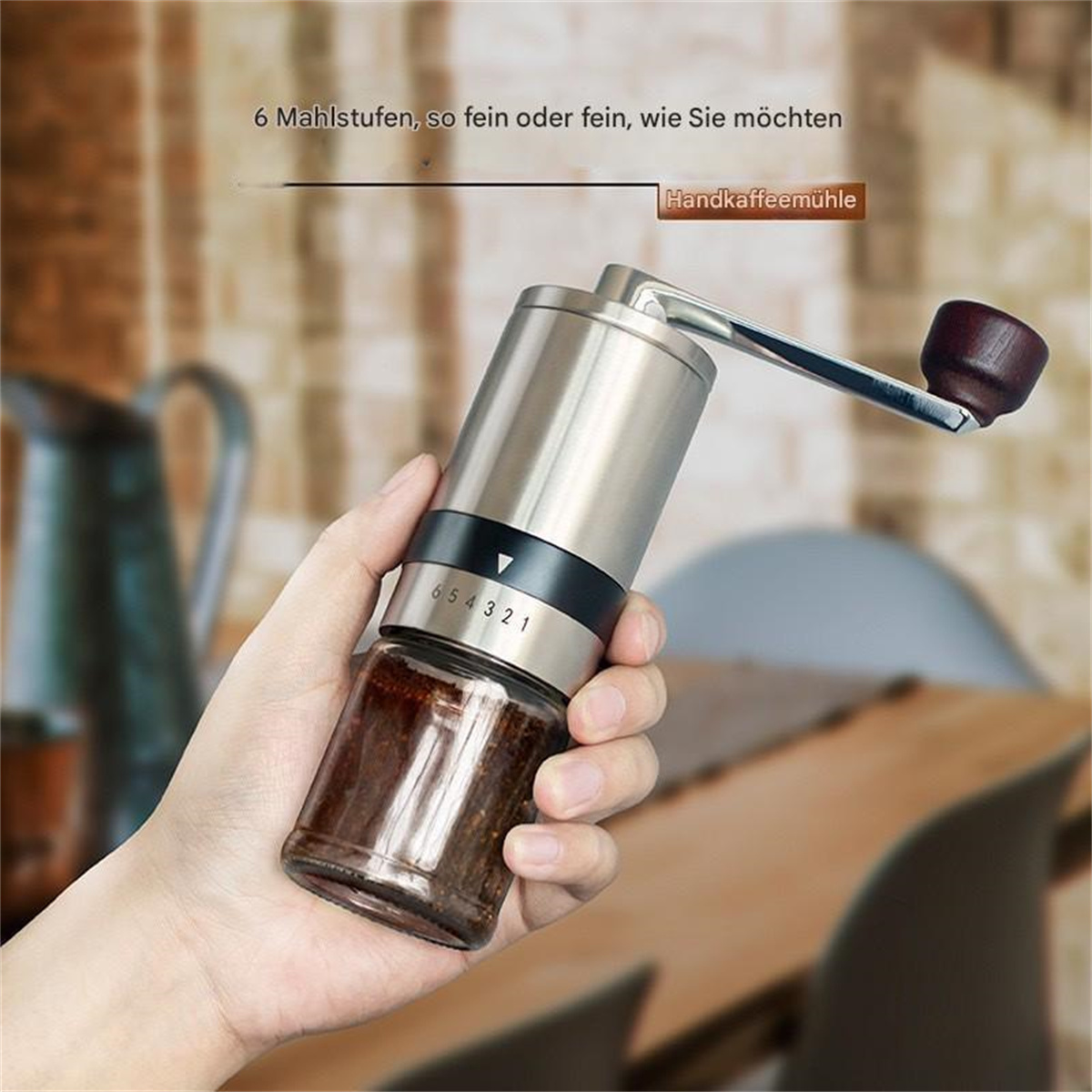 Eine Hand hält eine manuelle Kaffeemühle aus Edelstahl. Ein Glasbehälter enthält Kaffeepulver. Es hat 6 Mahlgradeinstellungen.