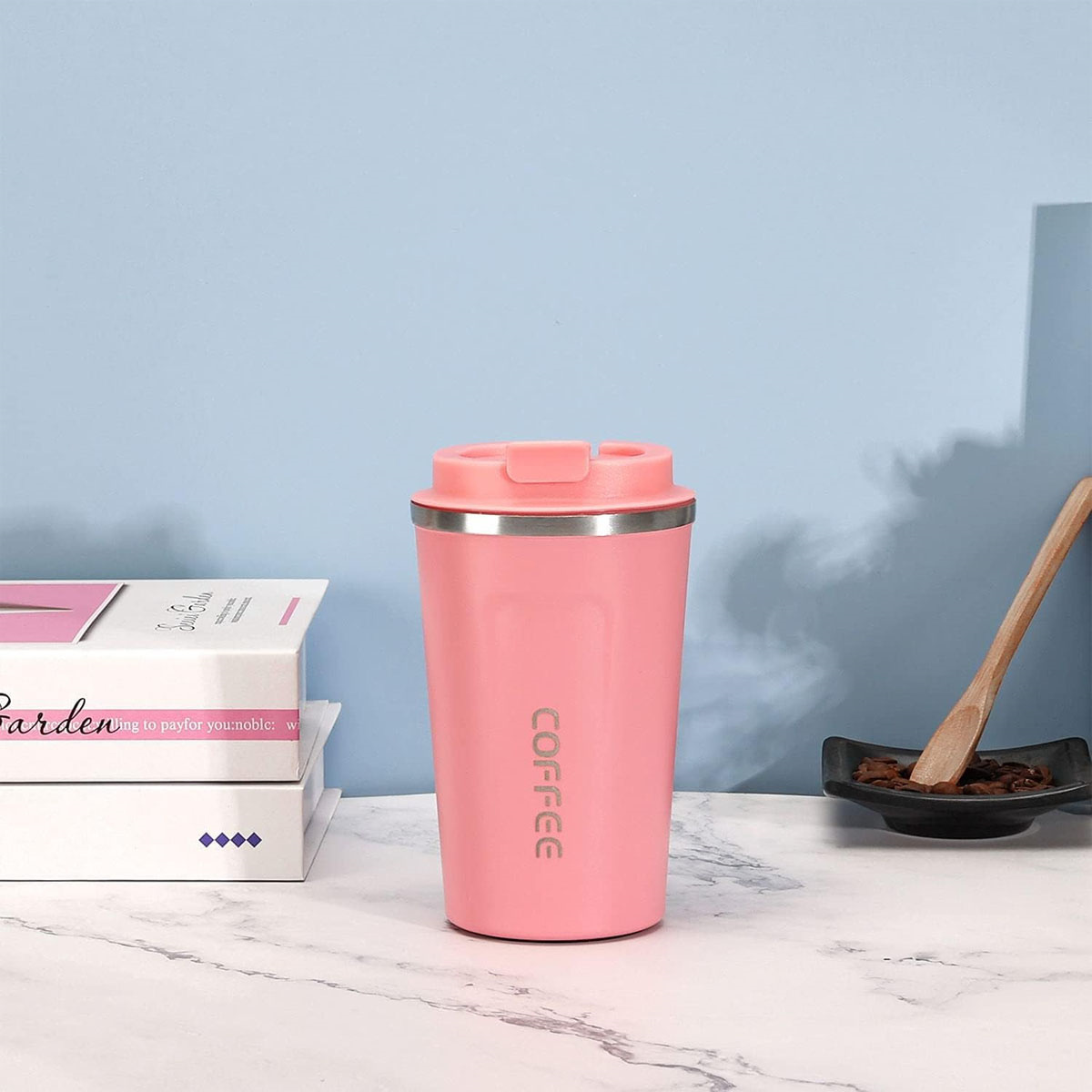 Rosa Reisebecher mit 'COFFEE'-Aufdruck. Steht neben Büchern und einem HolzLöffel.