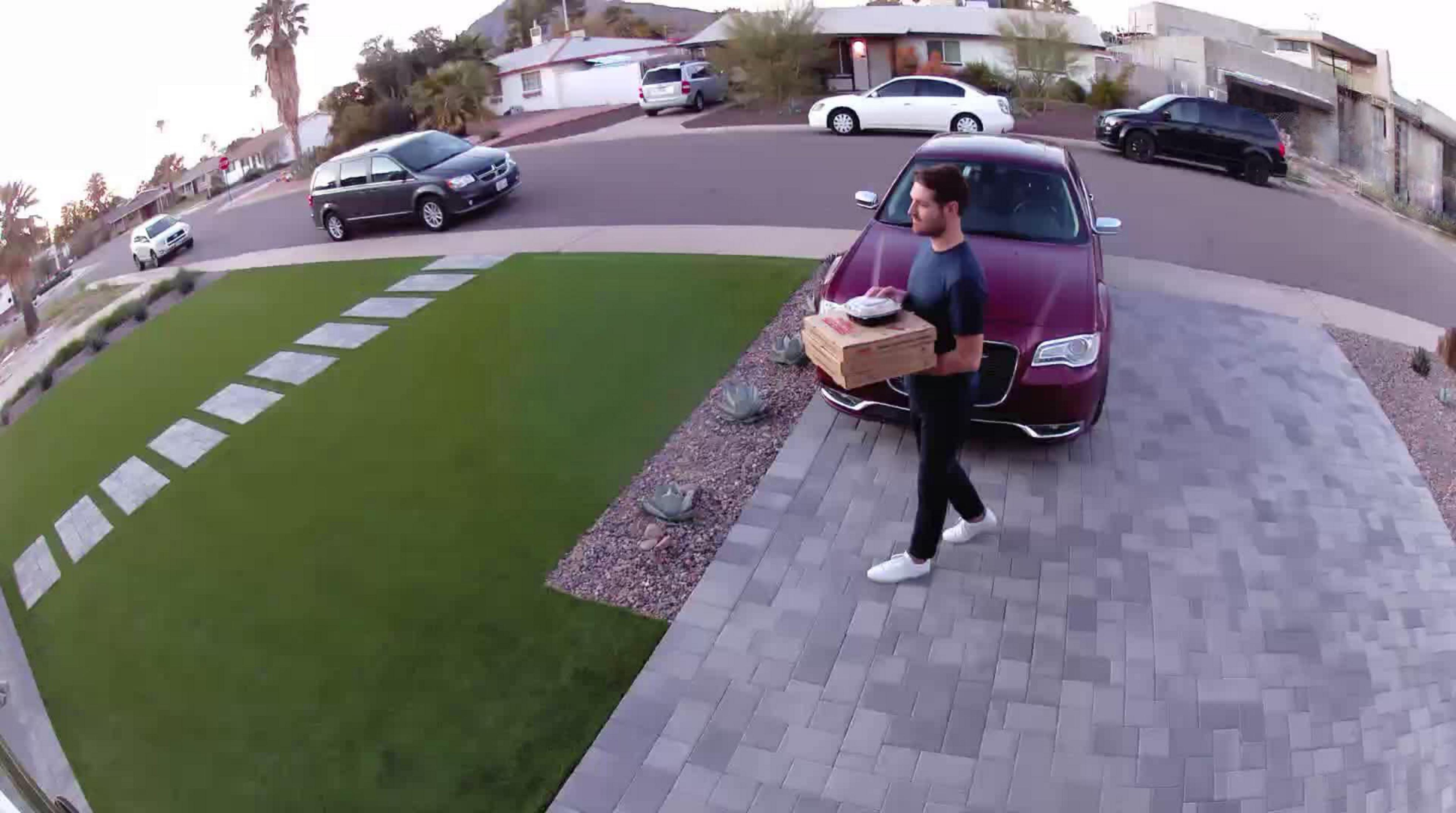 Mężczyzna niosący pudełko po pizzy idzie w kierunku czerwonego samochodu zaparkowanego przed domem.