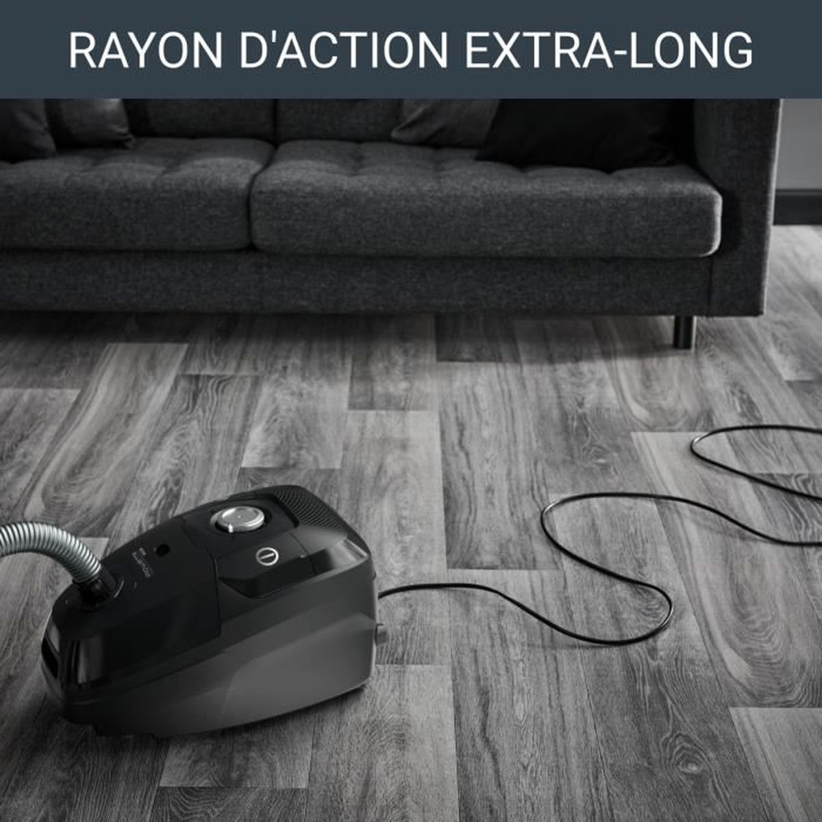 Czarny odkurzacz na szarej drewnianej podłodze, w tle szara sofa.