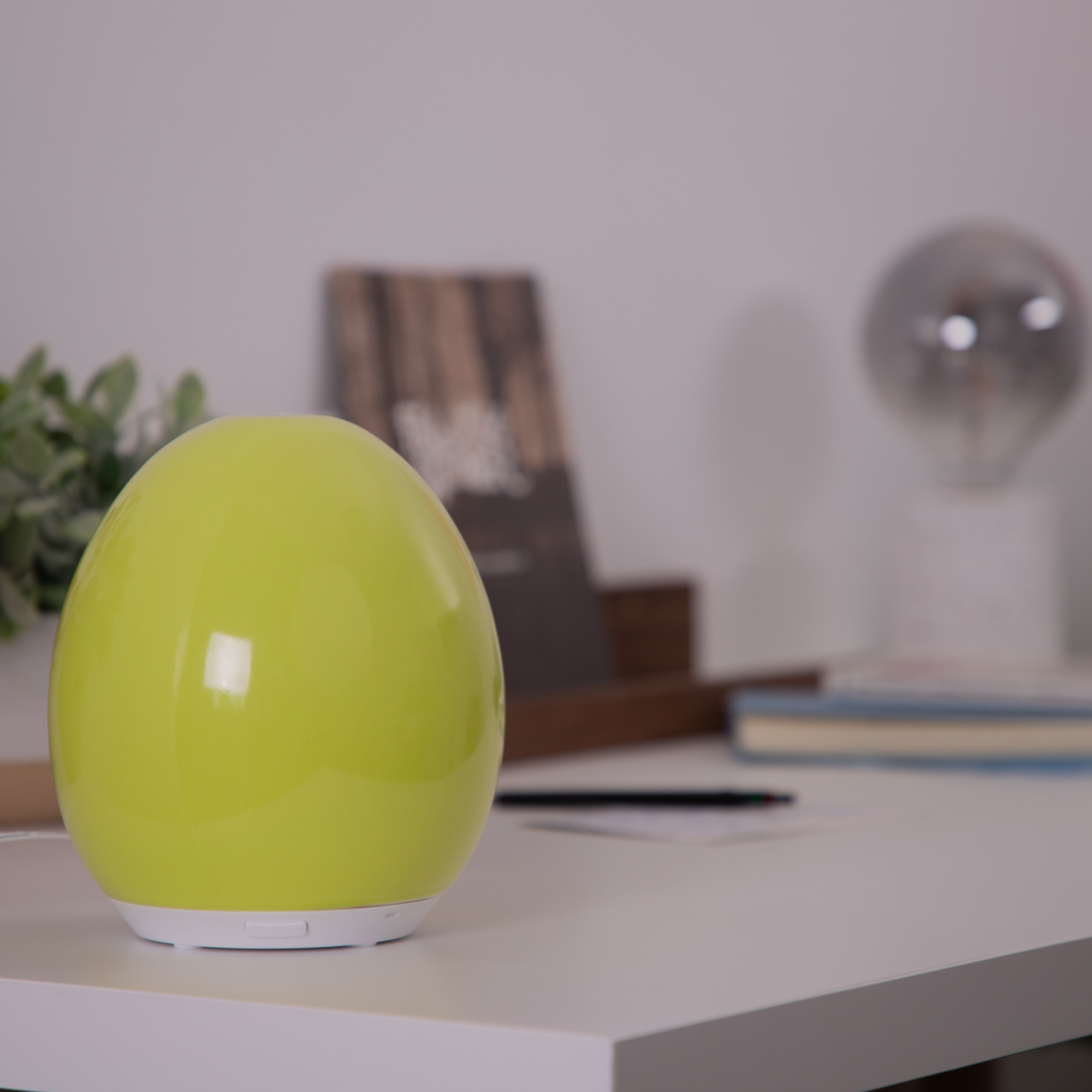 Un diffuseur vert en forme d'œuf est posé sur un bureau blanc. Un livre flou et une lumière sont en arrière-plan.