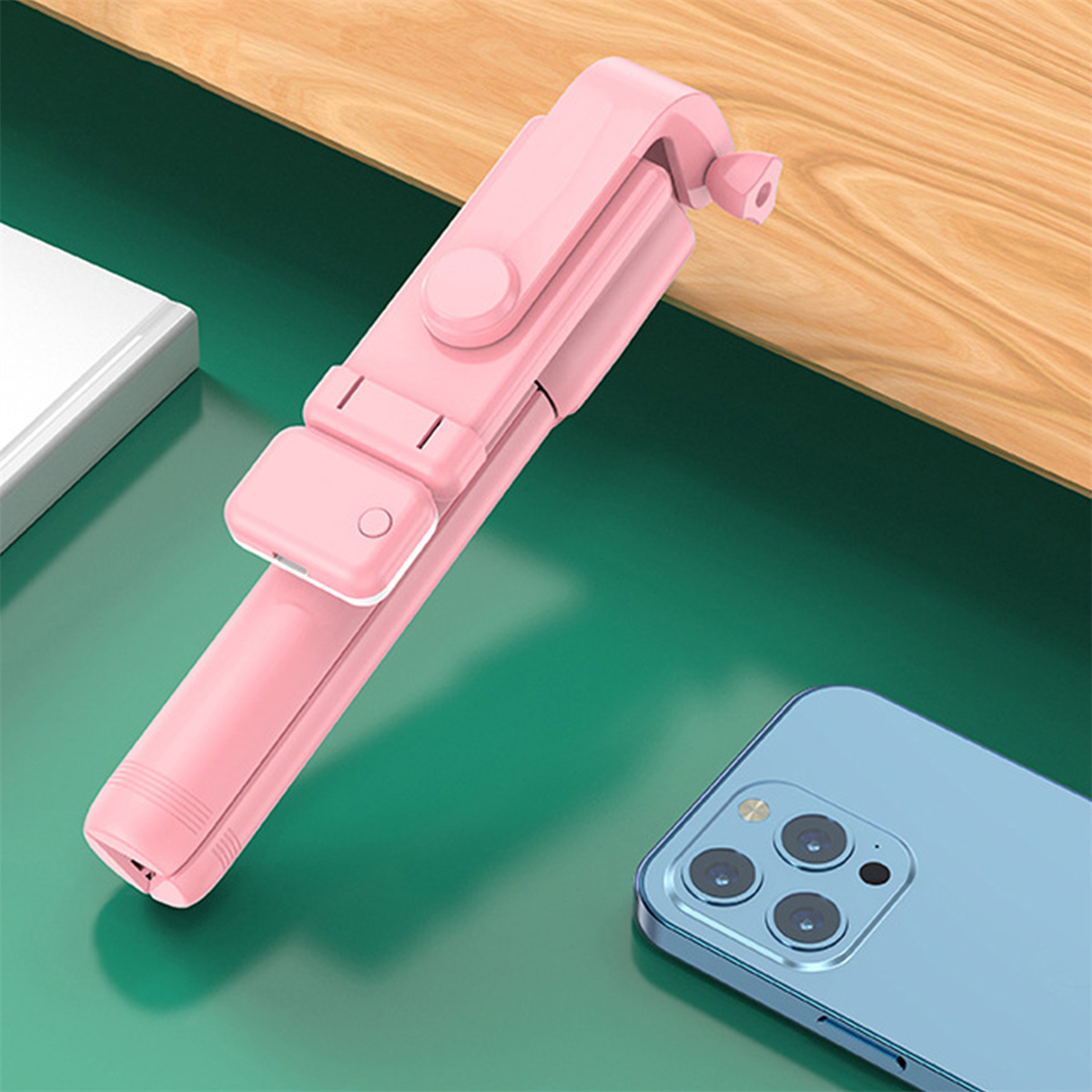 Pinkfarbener Selfie-Stick. Er ist zusammengeklappt und liegt neben einem blauen Telefon auf einer grünen Oberfläche.