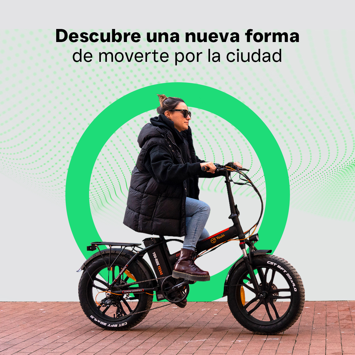 Kobieta na czarnym rowerze elektrycznym, w okularach przeciwsłonecznych, na brukowanej drodze, zielone koło.