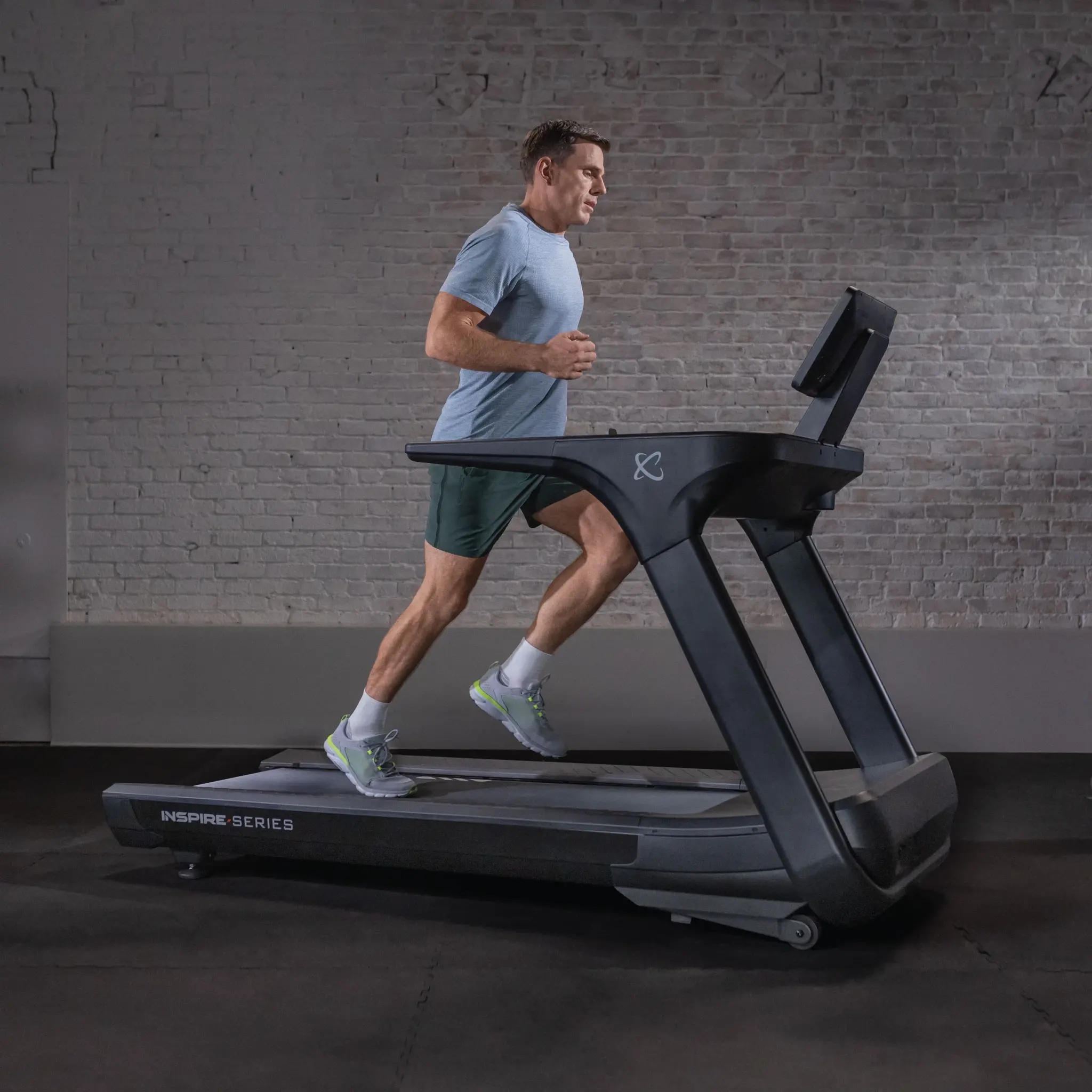 Man running on a black treadmill in a gym. He wears a blue shirt and green shorts.