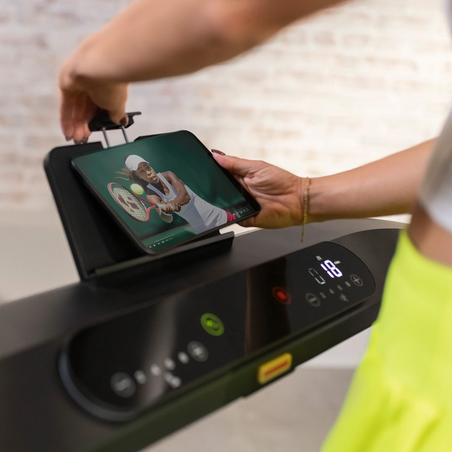 A person using a tablet to watch a tennis match while on a treadmill, with an 18 display.