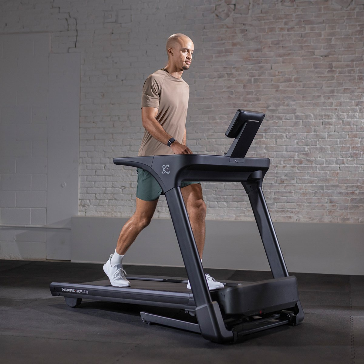 A man walks on a treadmill, with a brick wall background and a dark floor.