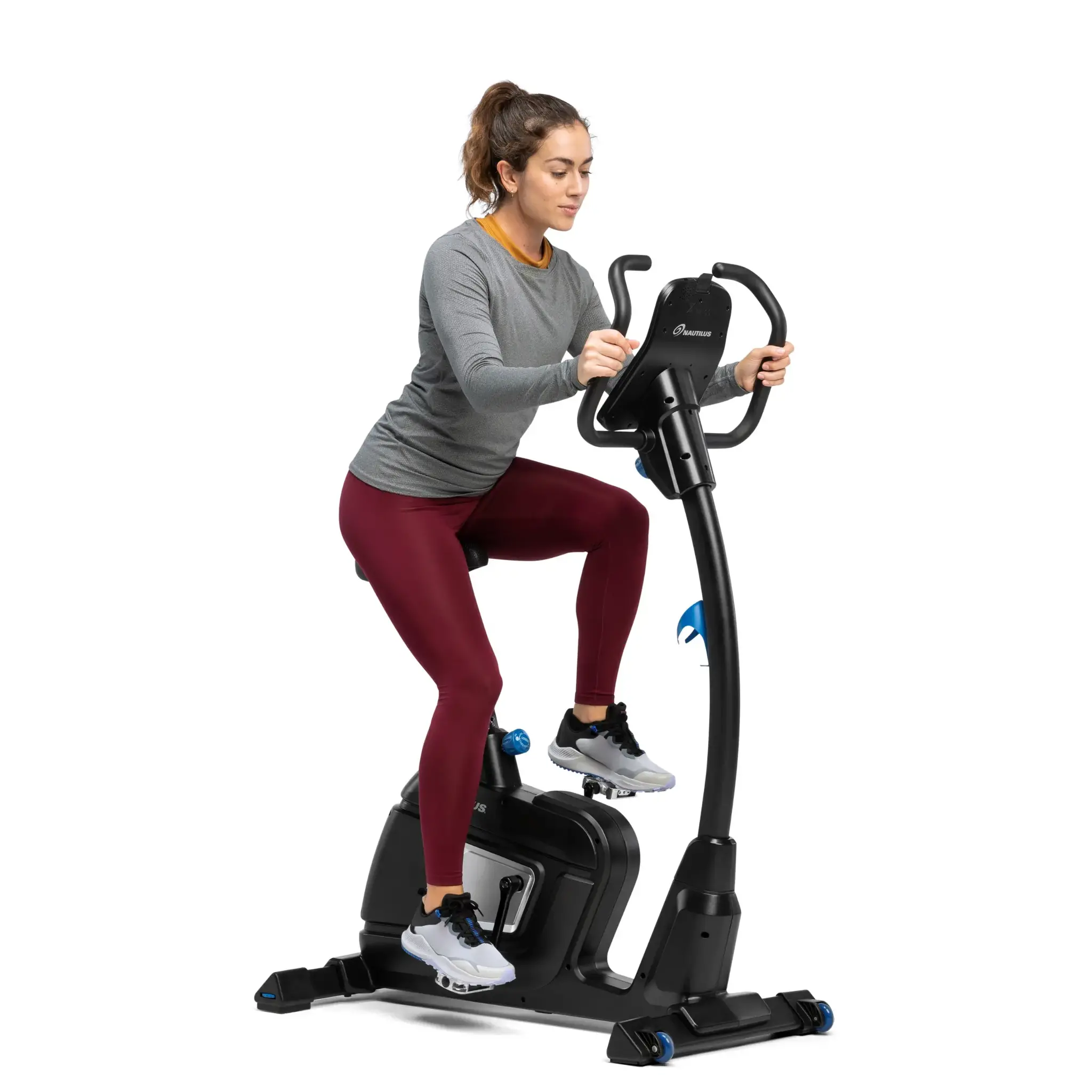 Frau trainiert auf einem Heimtrainer, trägt graues Hemd und rote Hose, in einem weißen Raum.