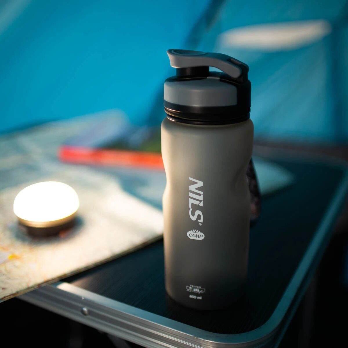 Eine graue Wasserflasche mit 'NILS' auf einem Tisch mit Licht und Karte.
