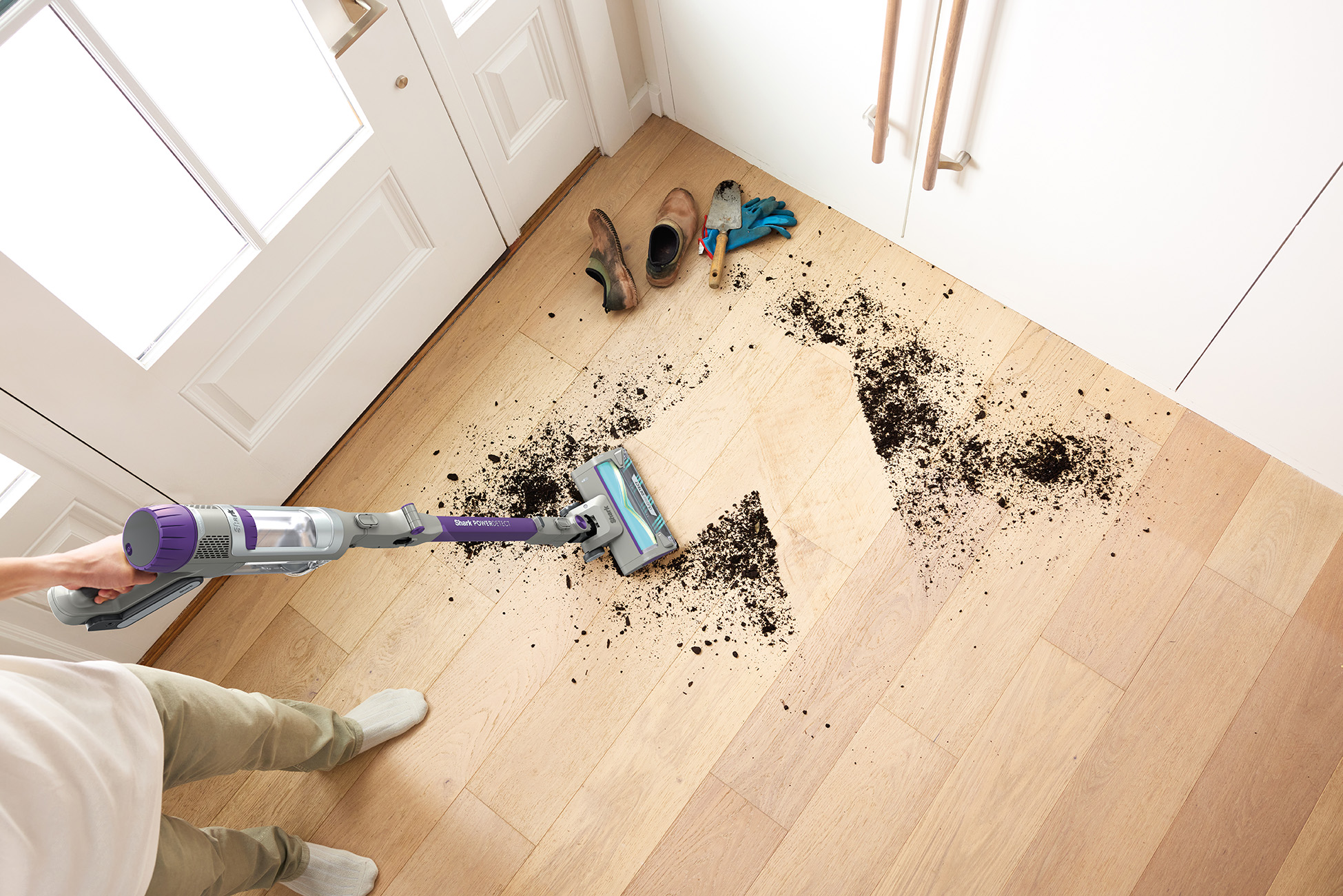 A person vacuums dirt on a wooden floor. Shoes and gloves are near a door.