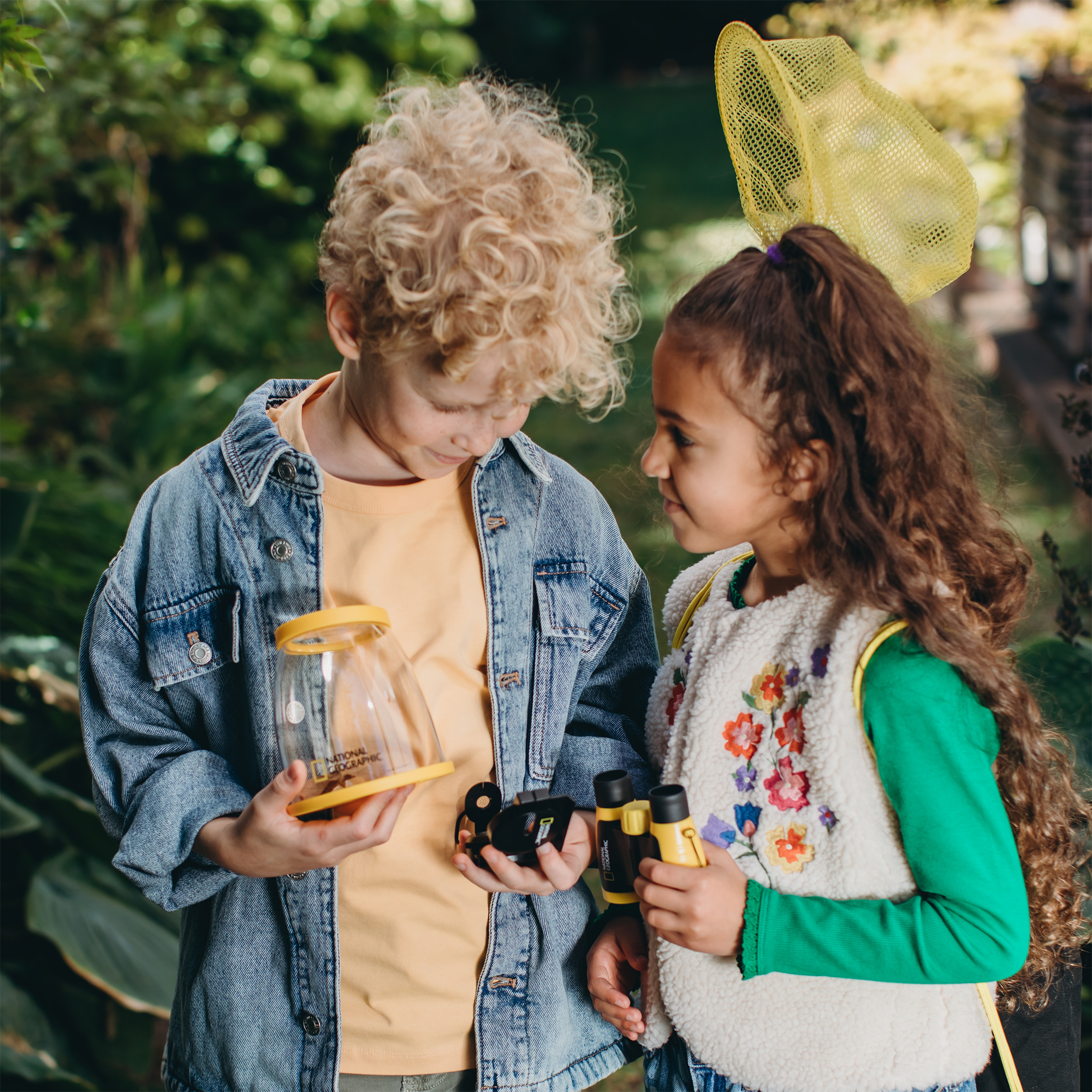 Due bambini esplorano la natura con un acchiappa-insetti, un binocolo e una rete.