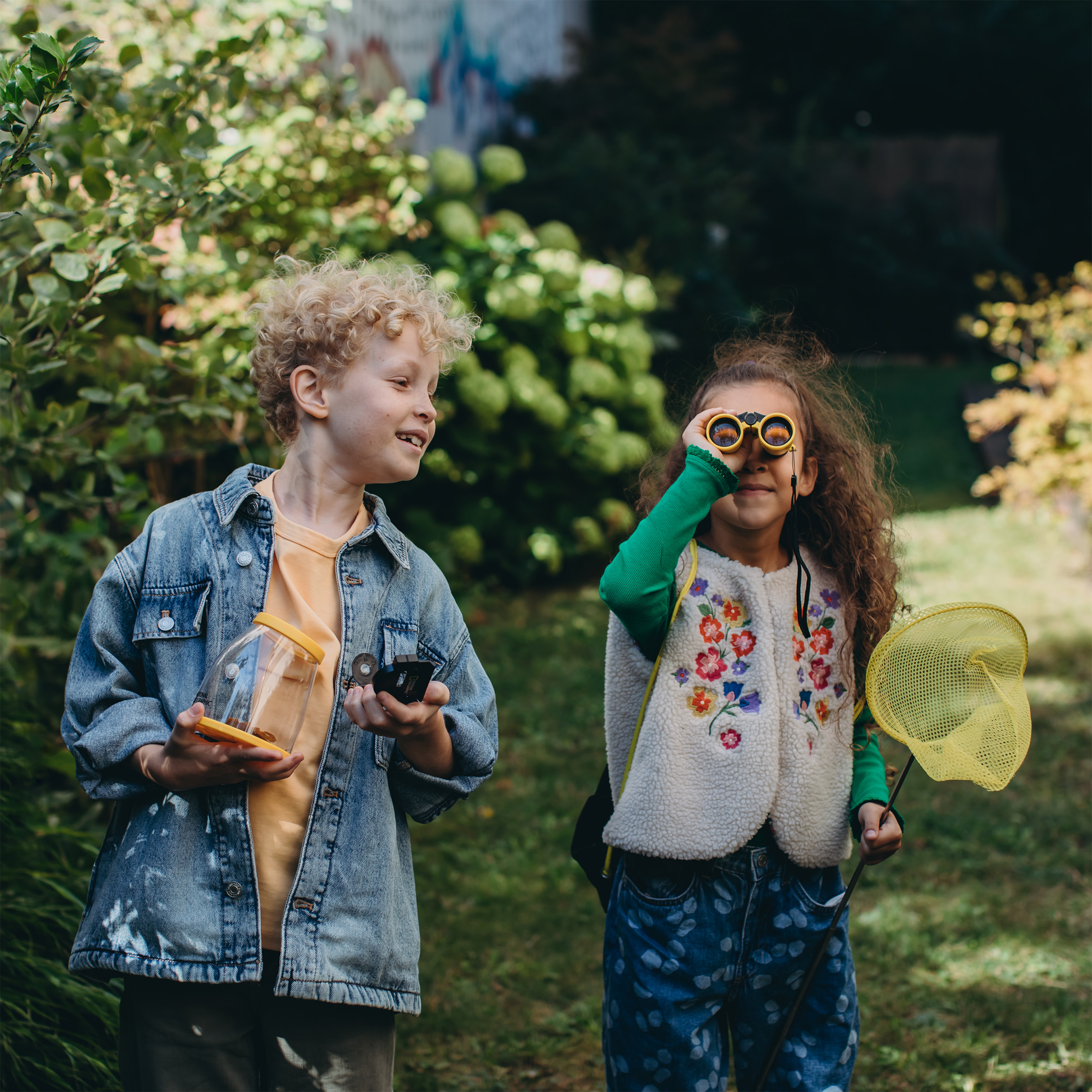 Due bambini in un giardino, uno guarda attraverso un binocolo, l'altro tiene un contenitore.