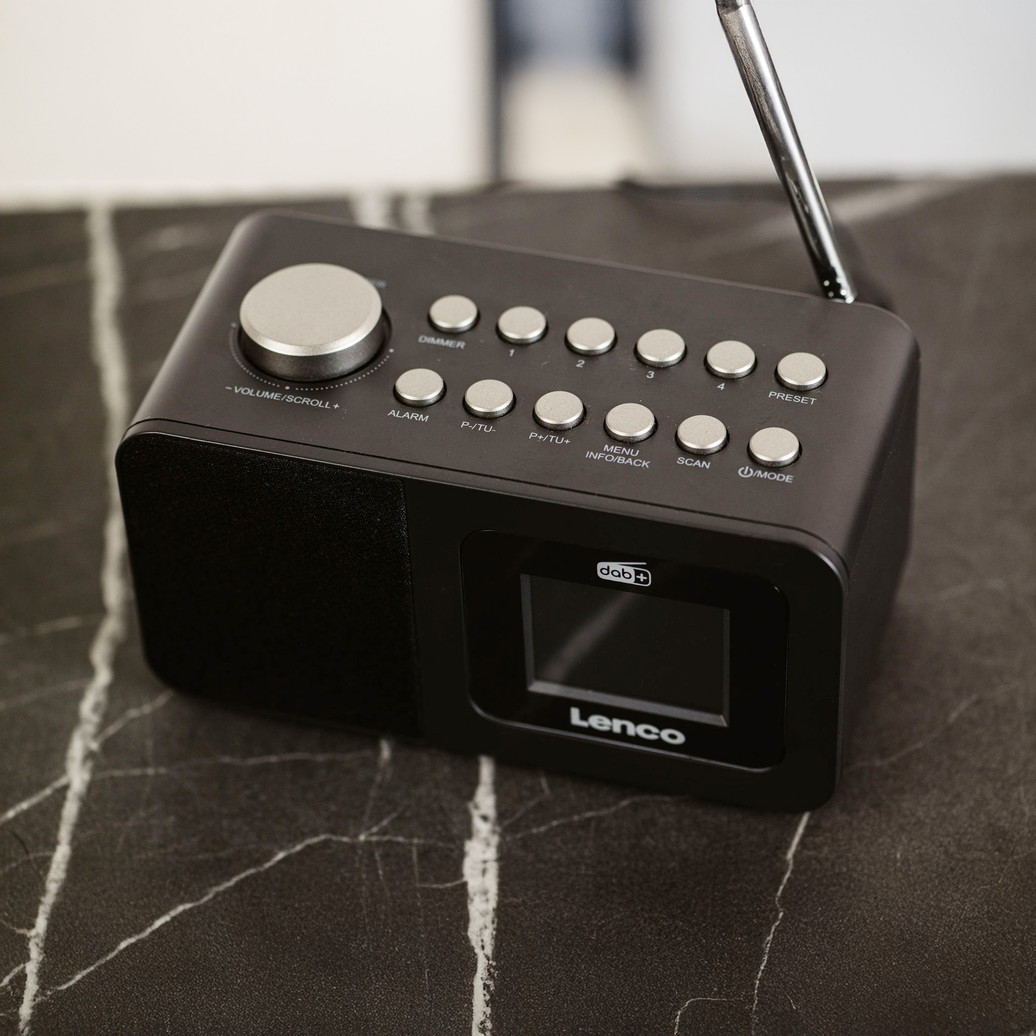 Czarny radioodbiornik DAB z przyciskami i wyświetlaczem, na marmurowej powierzchni. Antena wysunięta.