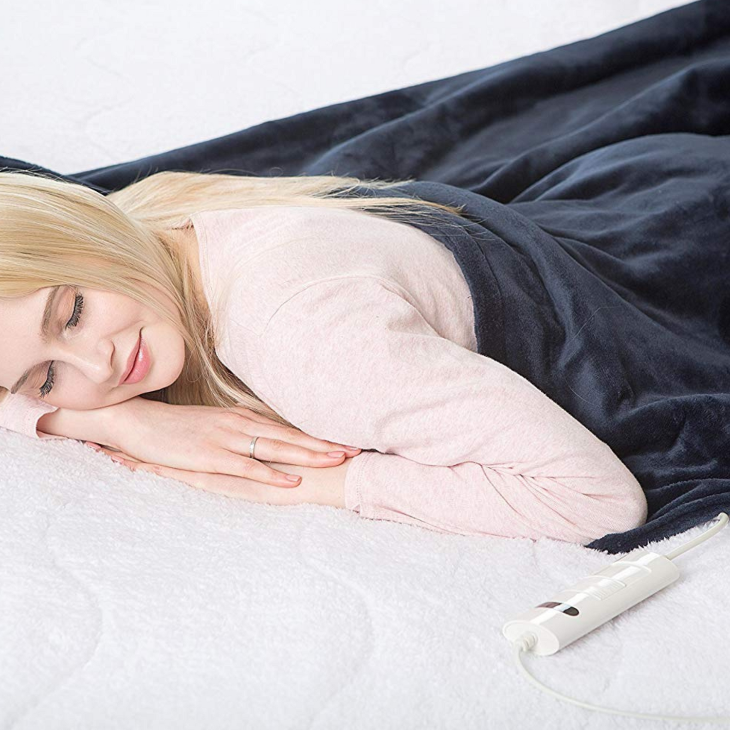 Mujer acostada en una cama, cubierta por una manta azul, con una camisa rosa. Dispositivo de control blanco visible.
