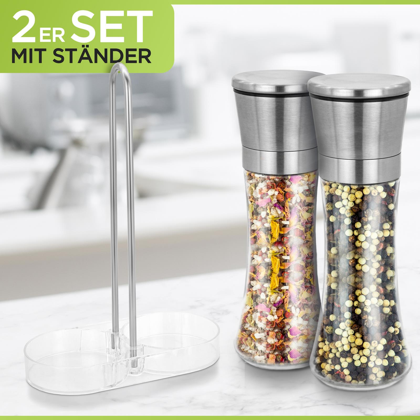 Zwei Glasmühlen mit Metallaufsätzen und einem Ständer, gefüllt mit Gewürzen, auf weißer Oberfläche.