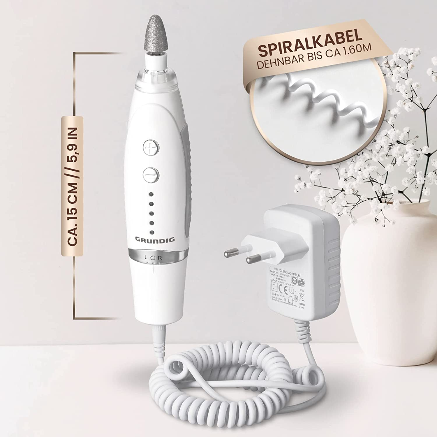 Ein weiß-grauer Grundig-Nagelfeile mit Netzteil und Spiralkabel auf weißer Oberfläche. Im Hintergrund eine Vase mit Blumen.