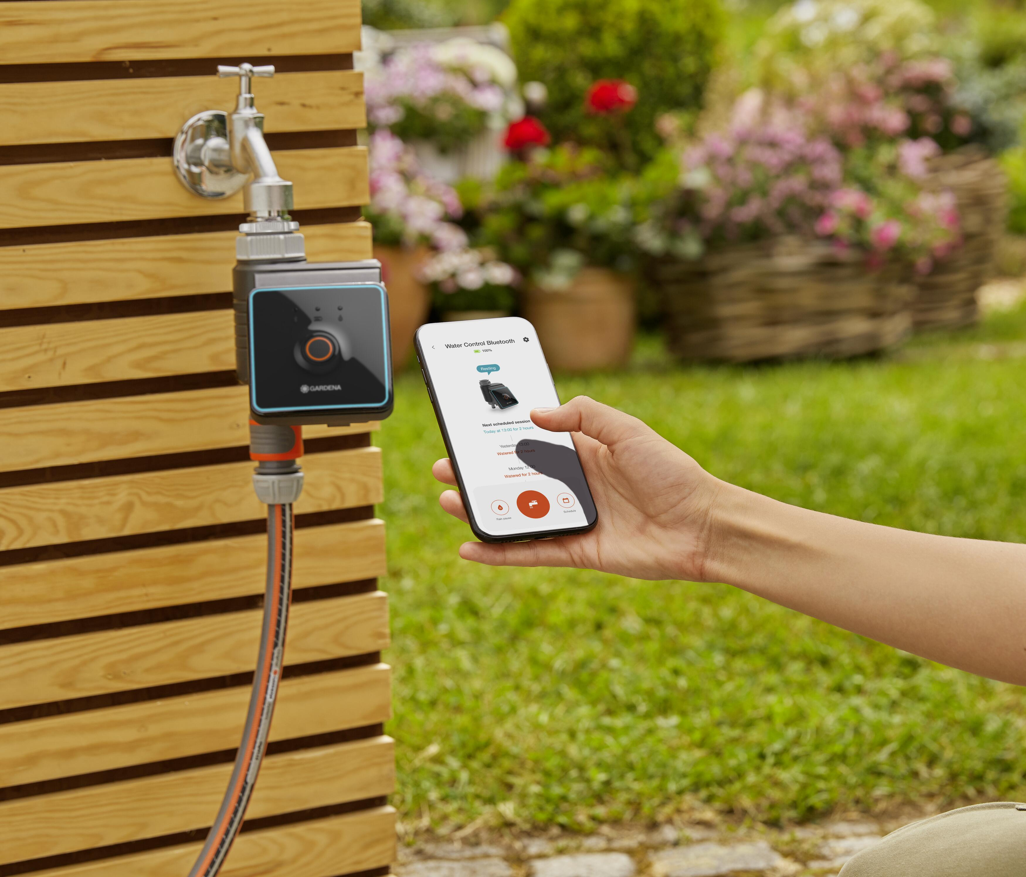Eine Person steuert mit einem Smartphone ein Wasserventil, das an einem Holzzaun angeschlossen ist.