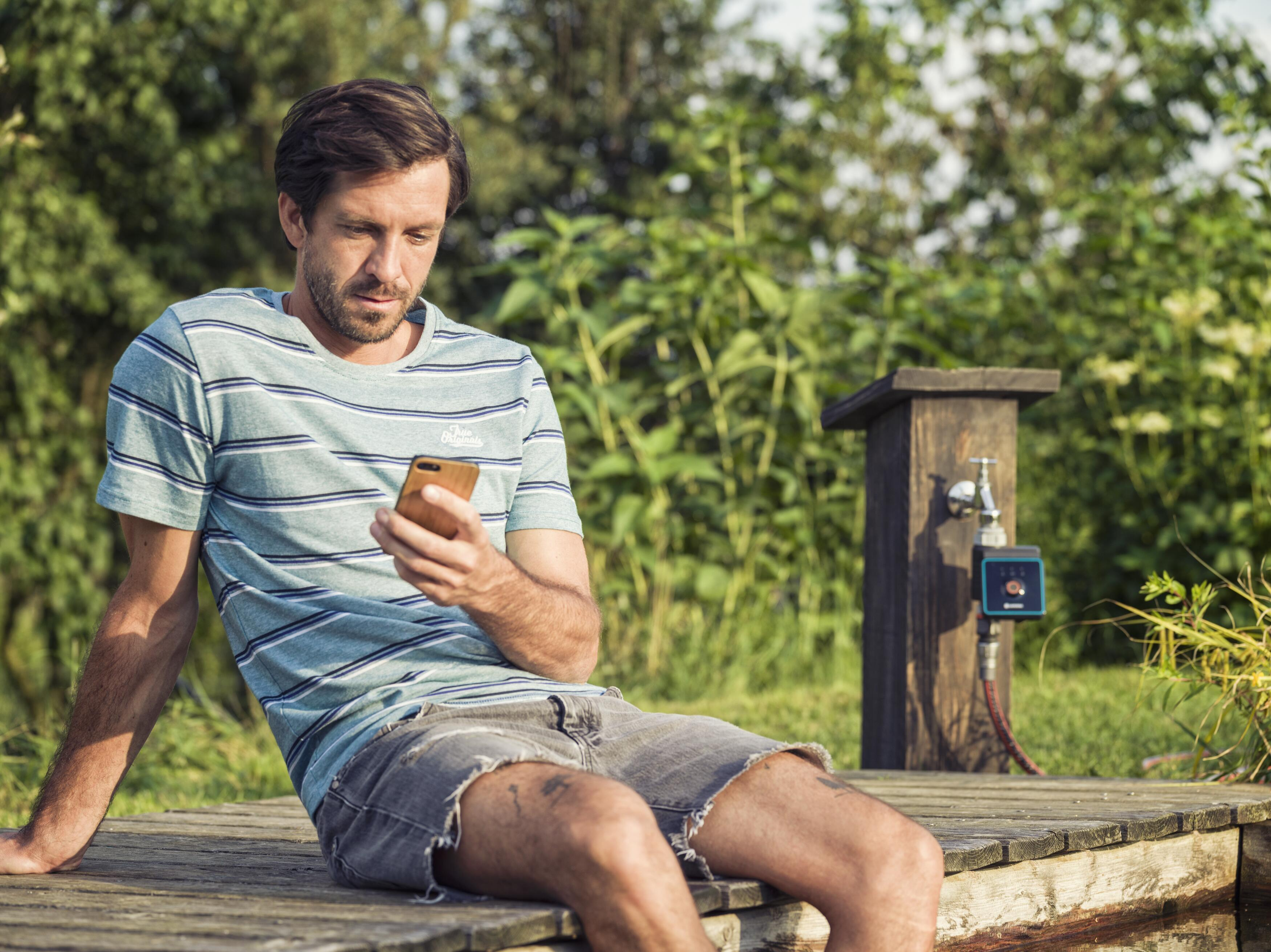 Ein Mann sitzt auf einem Holzsteg und schaut auf sein Handy. Hinter ihm grünes Laub und eine Wasserhahn-Konstruktion.