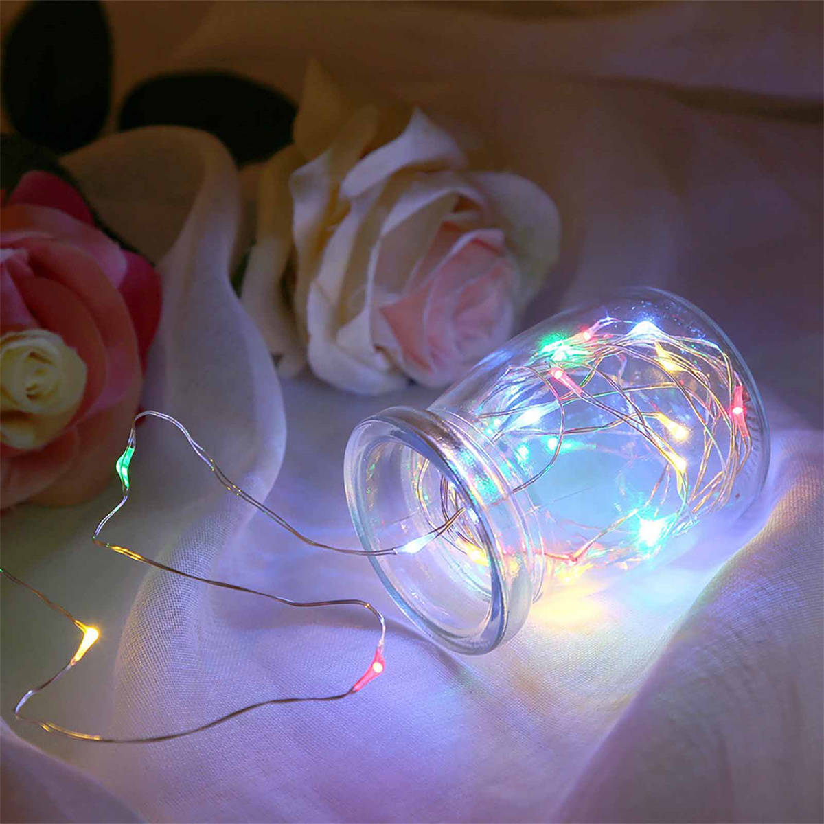 Ein Glasgefäß mit bunten Lichterketten. Rosen im Hintergrund. Schafft eine romantische Stimmung.