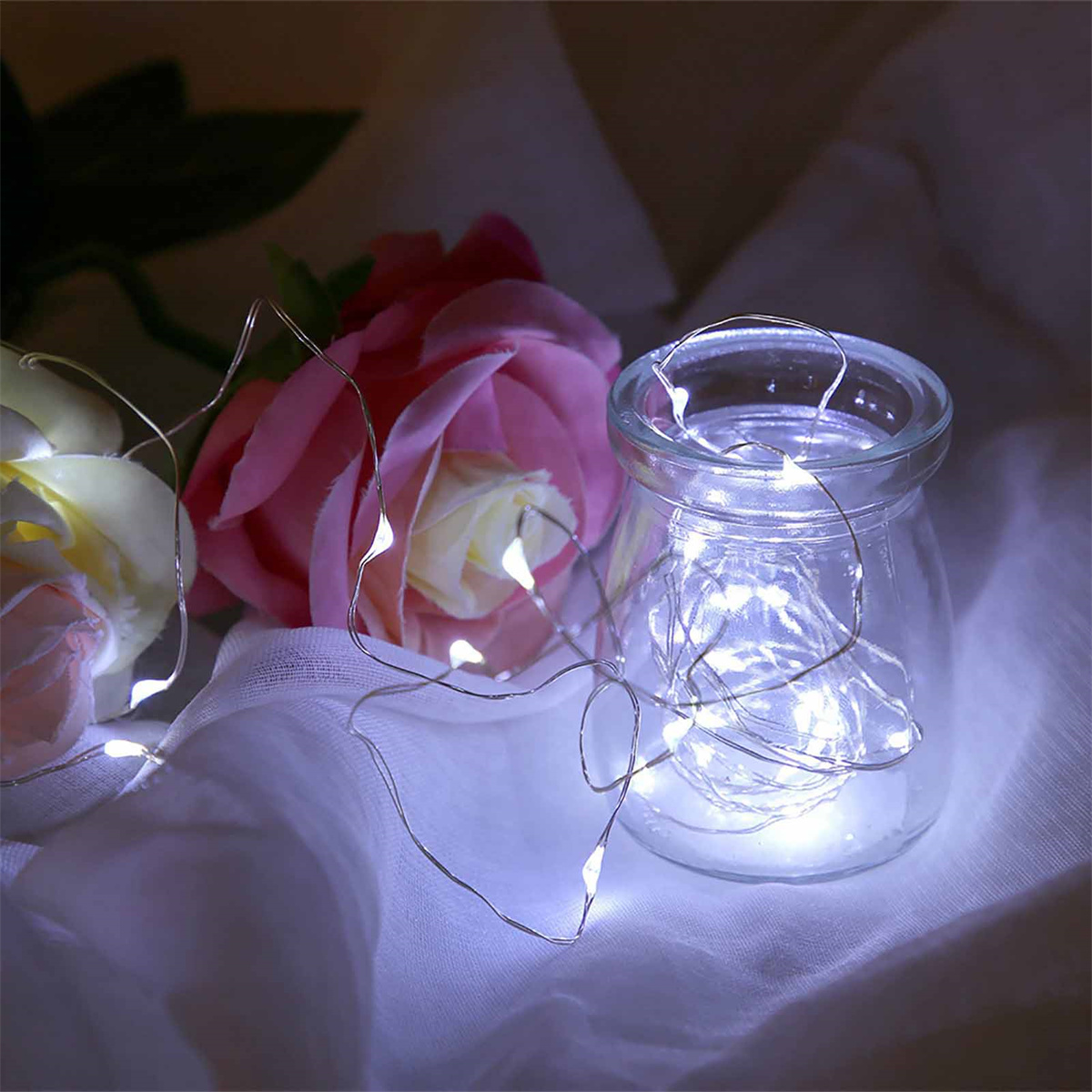 Ein Glasgefäß gefüllt mit kleinen weißen LED-Lichtern, mit rosafarbenen Rosen und weißem Stoff im Hintergrund.