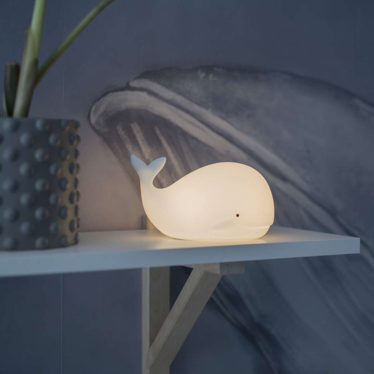 Eine walförmige Lampe steht auf einem weißen Regal. Es gibt eine graue Wand mit einer Walzeichnung.