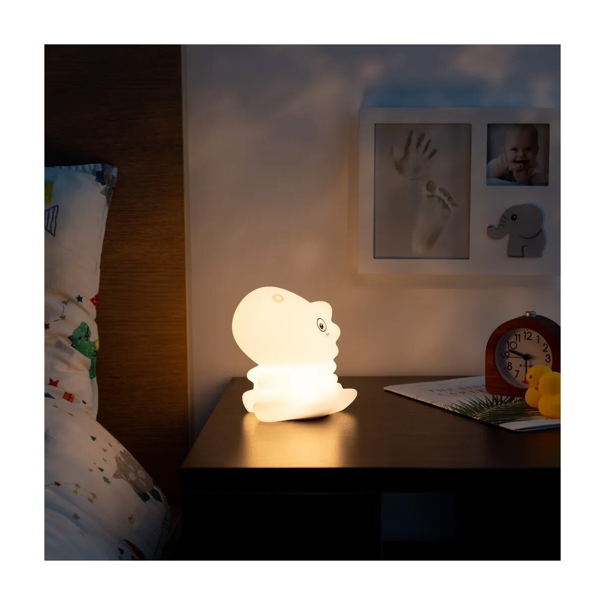 Ein leuchtendes weißes Dinosaurier-Nachtlicht auf einem Nachttisch. Dahinter ist ein gerahmtes Foto.