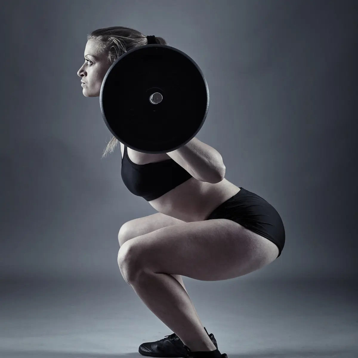 Woman in black activewear squats with a barbell on her back against a gray background.