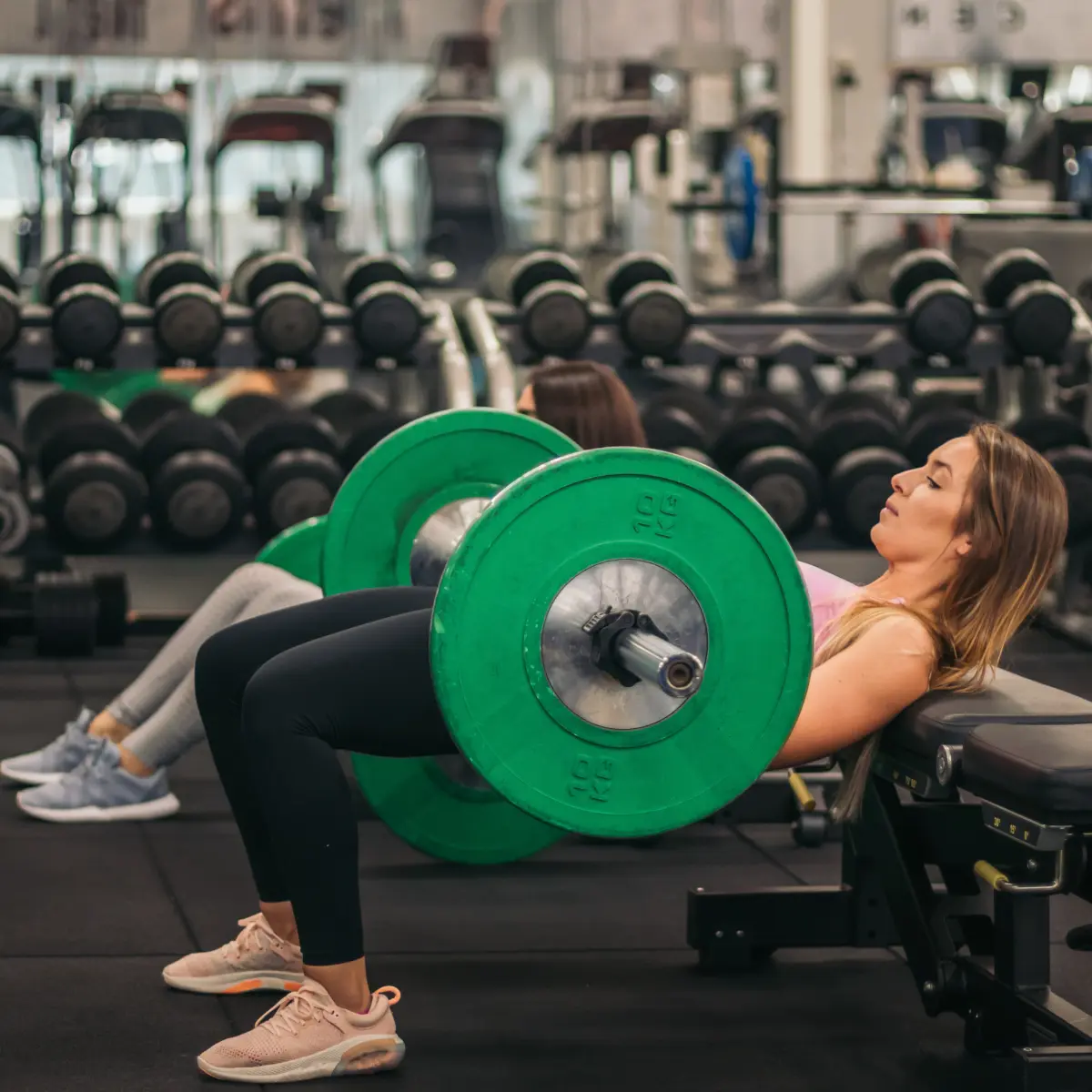 Two women exercising in a gym, using a barbell and bench.