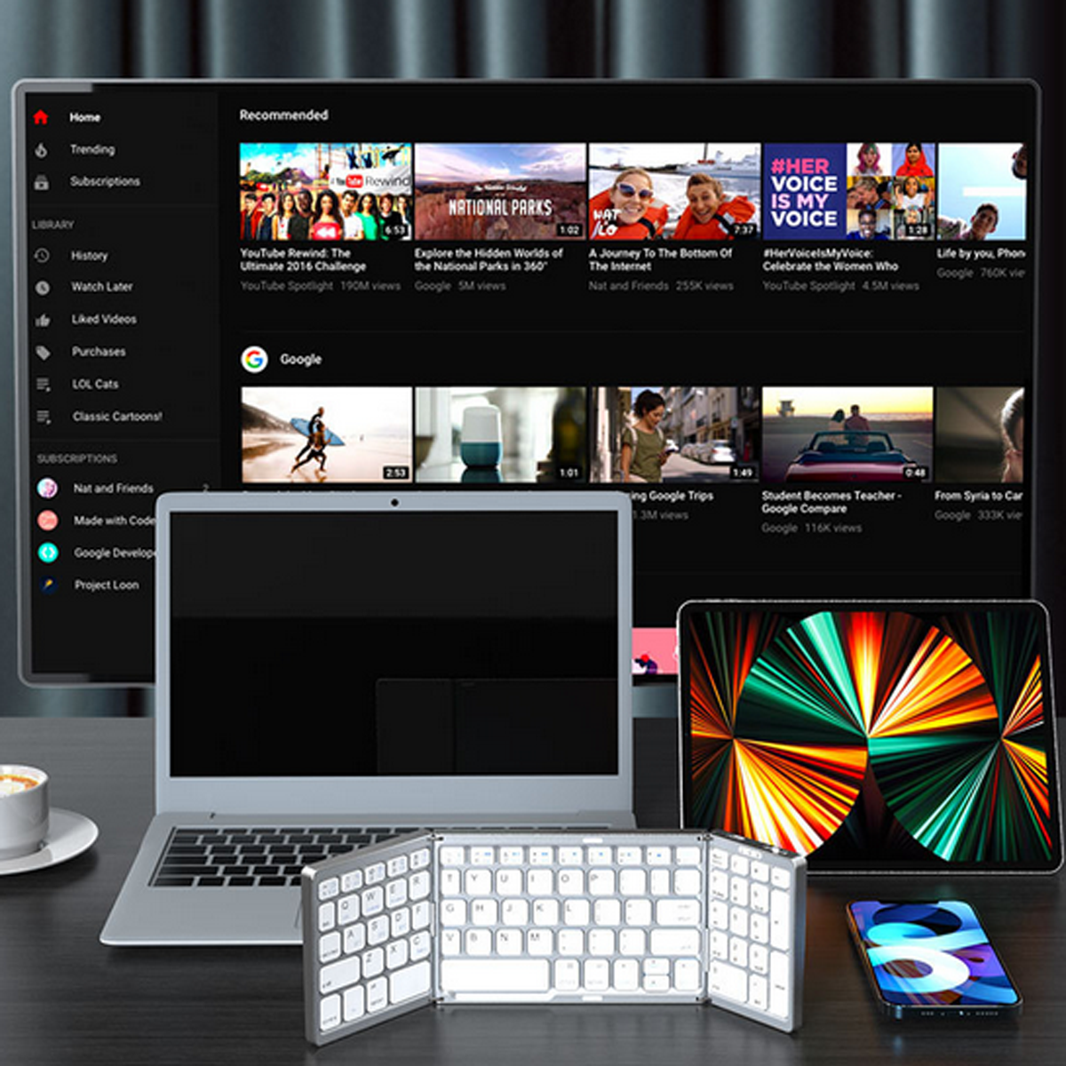 Ein Laptop, Tablet und eine gefaltete Tastatur auf einem Schreibtisch. Ein großer Bildschirm mit YouTube-Videos dahinter.