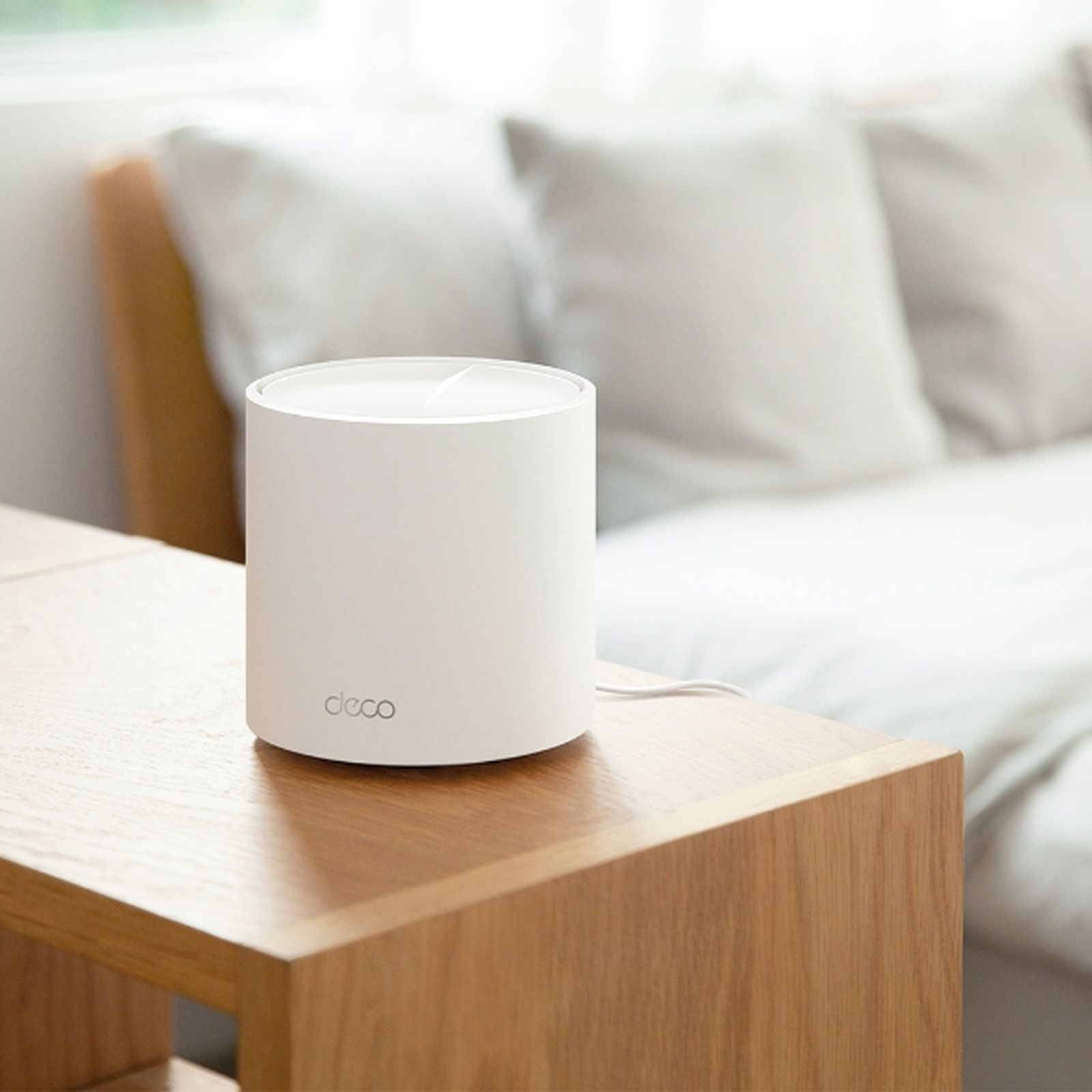 Biały cylindryczny router na drewnianym stoliku, obok łóżka z poduszkami w tle.