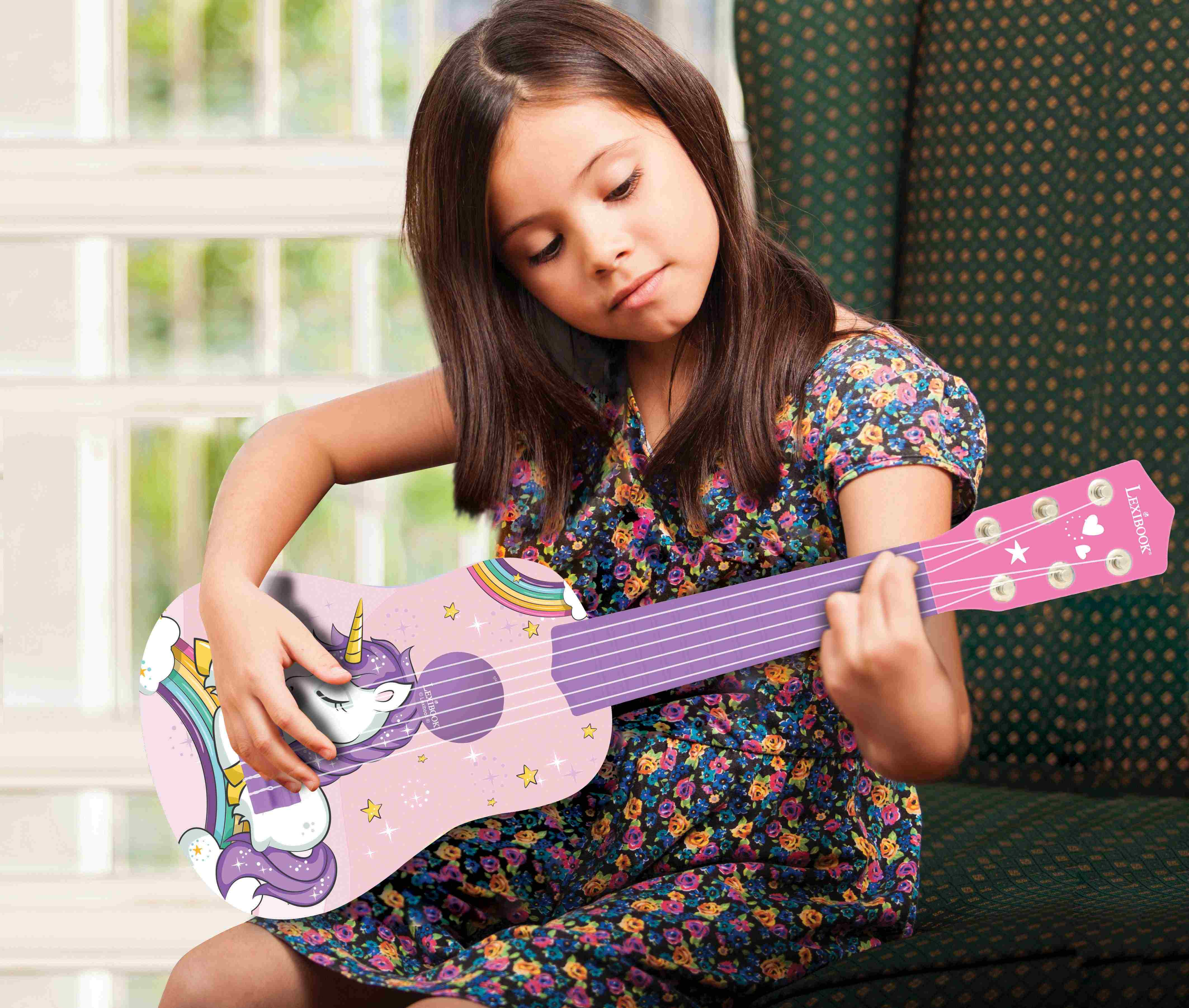 Una niña tocando un ukelele rosa con diseño de unicornio, sentada en un sofá floral.