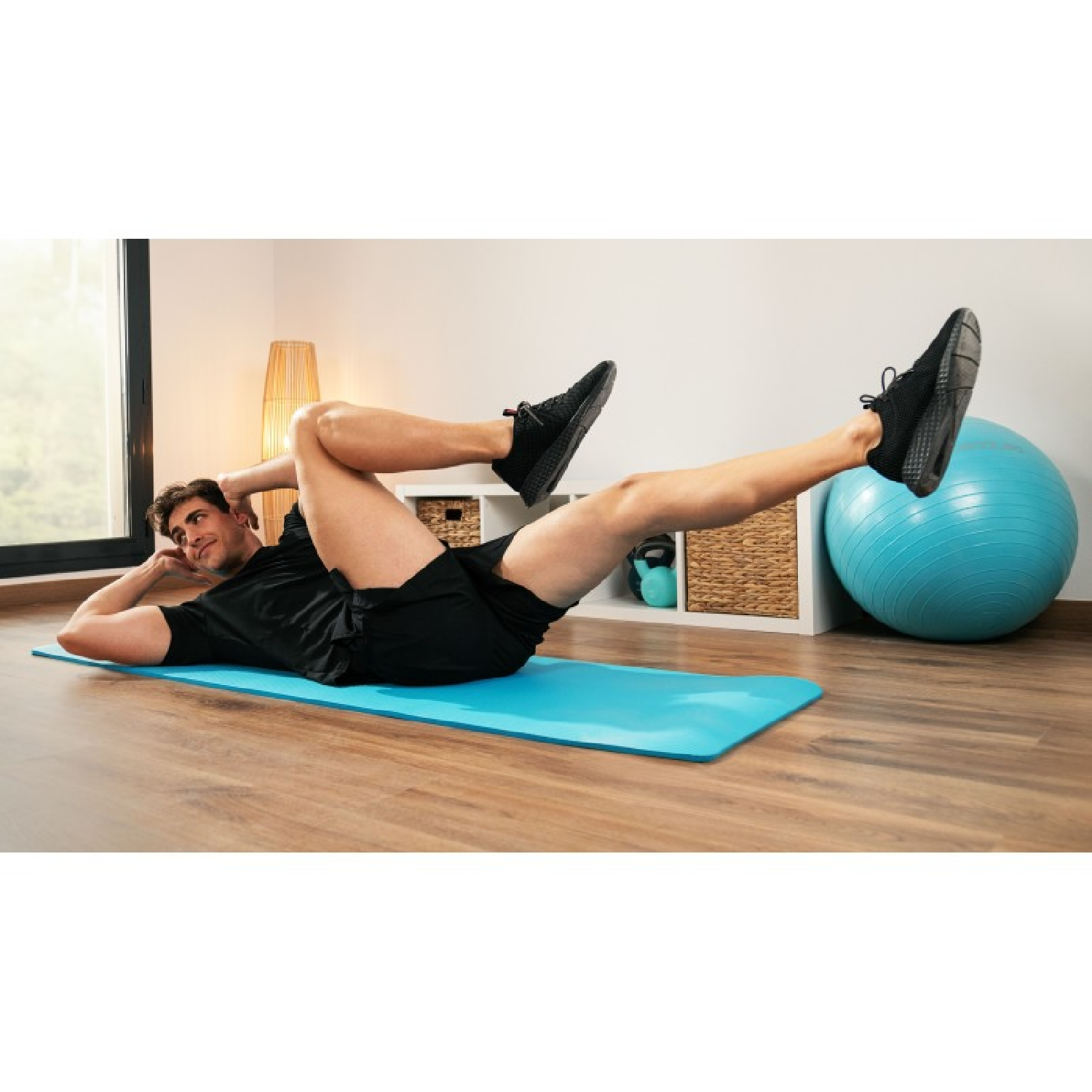 Uomo su tappetino blu fa crunch. Pantaloncini neri, scarpe nere. Palla yoga e finestra sullo sfondo.