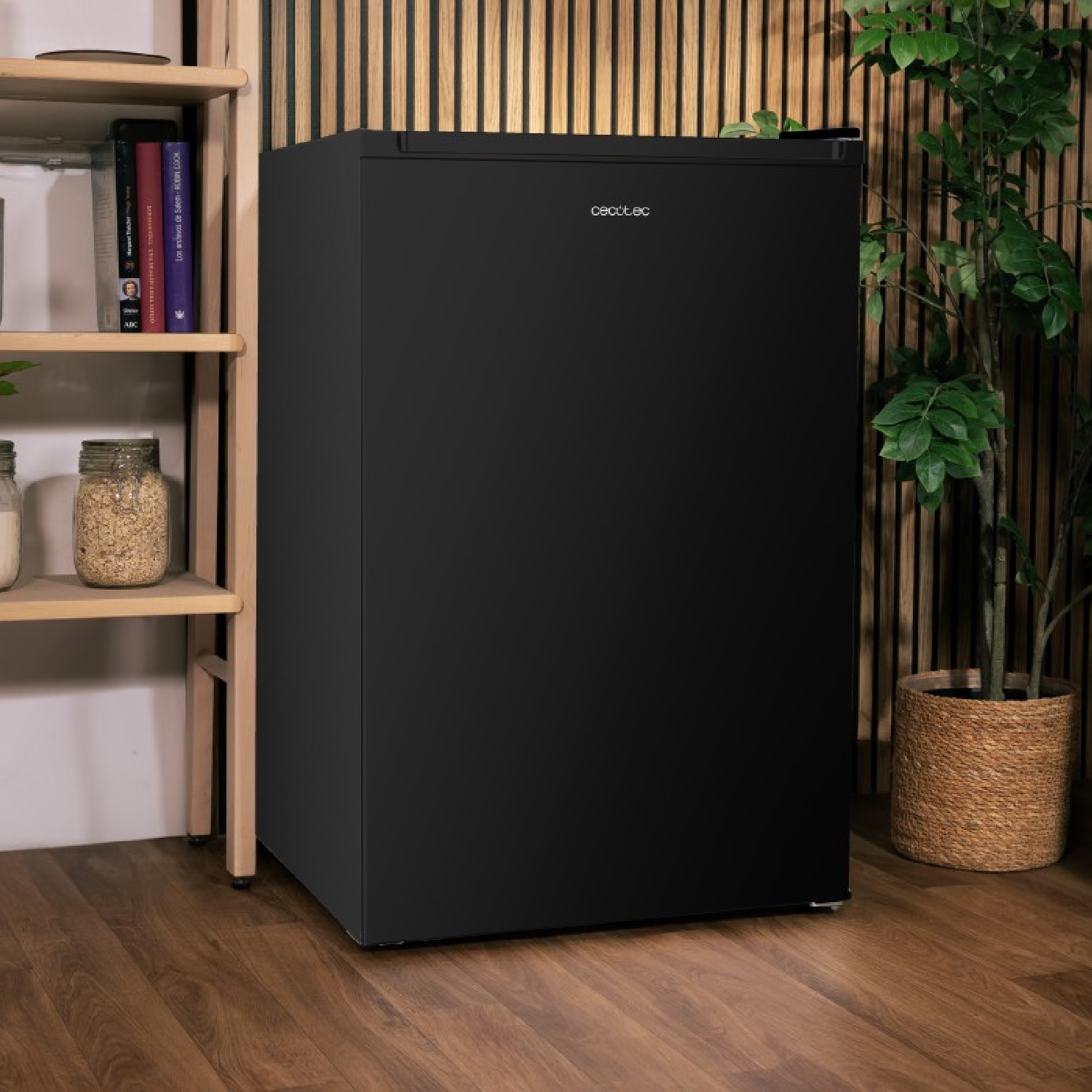 Un mini frigo nero in piedi su un pavimento di legno, con una mensola di legno e una pianta nelle vicinanze.