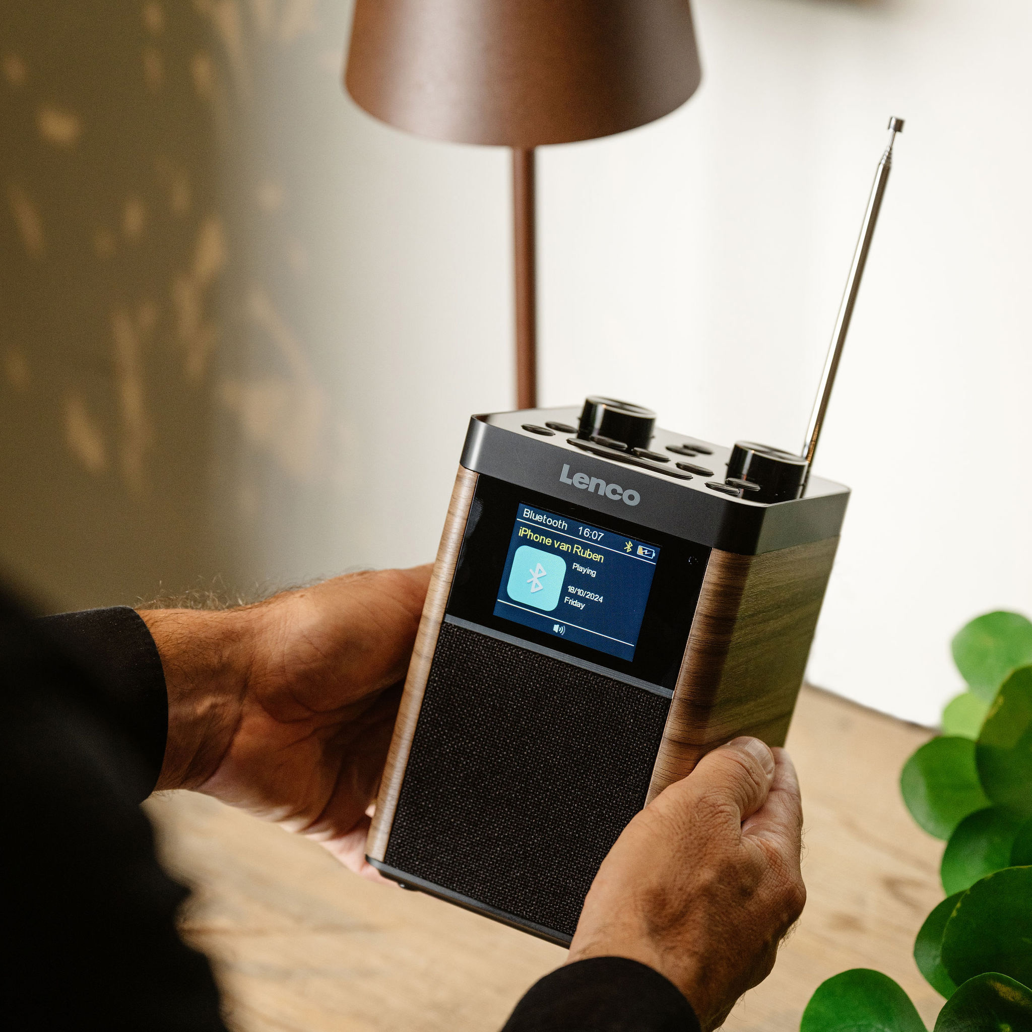 A person holds a wooden Lenco radio. The radio displays a screen with information. A lamp is in the background.